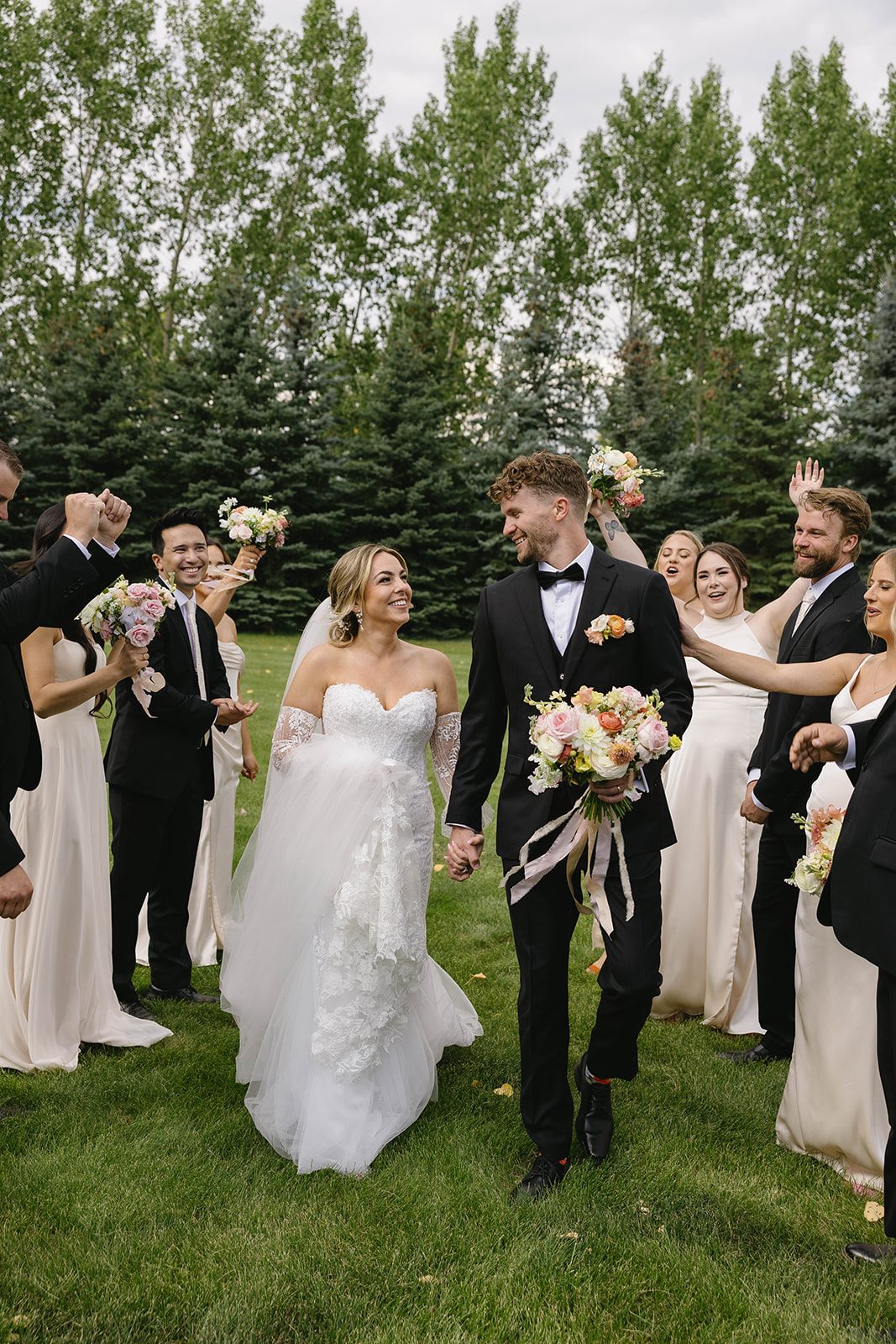Bride and groom surrounded by their celebrating wedding party look happily at each other, holding hands while the groom carries her pastel garden inspired bouquet of dahlias, garden roses, stock, lisianthus, ranunculus, sweet peas and orlaya.