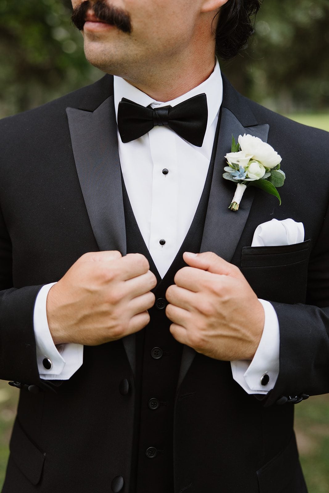 Cassidy + Adon Picture of the groom from the mouth down, holding onto the lapels of his black suit with an elegant white boutonnière featuring Ranunculus, spray ross and accented with tweedia.