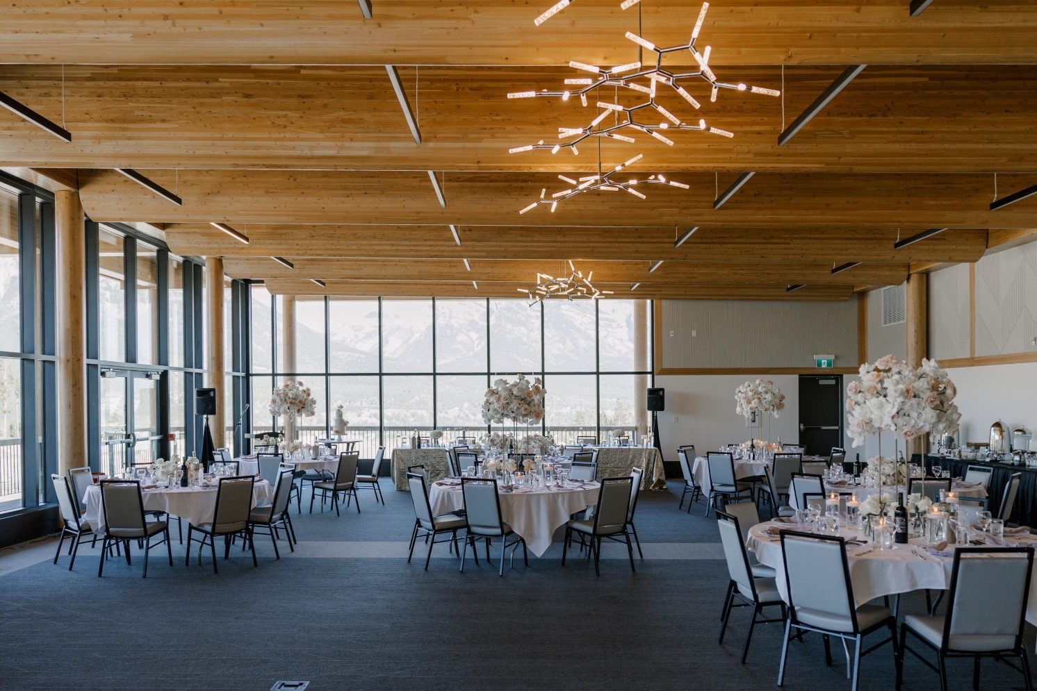 Reception Set Up Wide view of luxury wedding reception with tall floral centrepieces, modern lighting, and floor to ceiling windows showcasing the gorgeous mountain views.