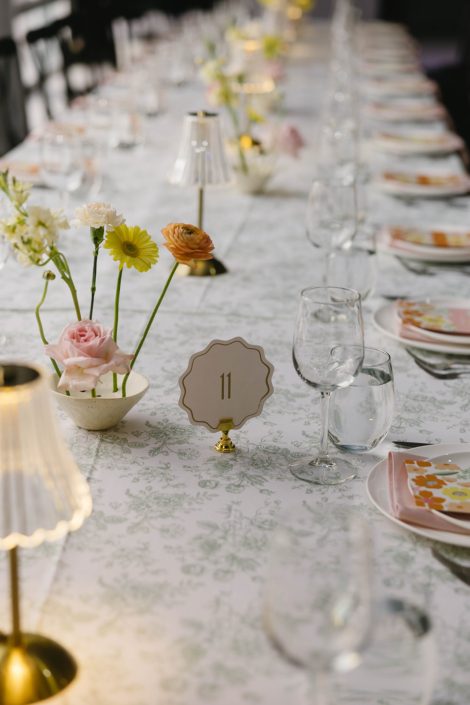 Close up of ikebana centrepiece with a pink rose, ranunculus, yellow gerbera daisy, and butter coloured stock on softly patterned ivory table linens, with striped napkins and floral stationery at each place setting.