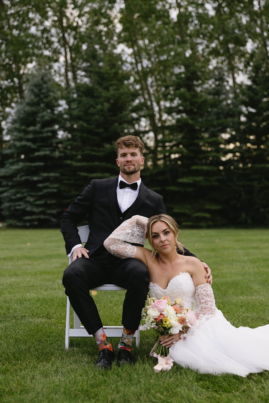 Bride and groom sit in the grass of Tangled Roots in Edmonton, with the tree line behind them. Bride holds a pastel, garden inspired bouquet of dahlias, garden roses, stock, lisianthus, ranunculus, sweet peas, and orlaya.
