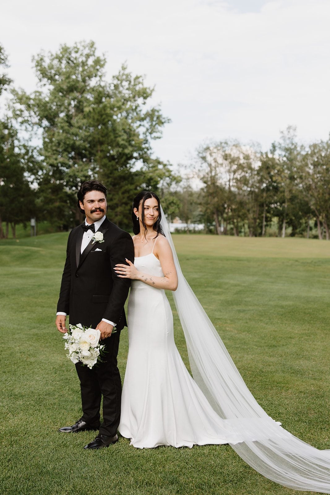 Cassidy + Adon Bride and groom standing side by side on the beautiful grounds at hilltop. Bride is resting her hand on the grooms arm, and he is holding her white bridal bouquet casually at his side.