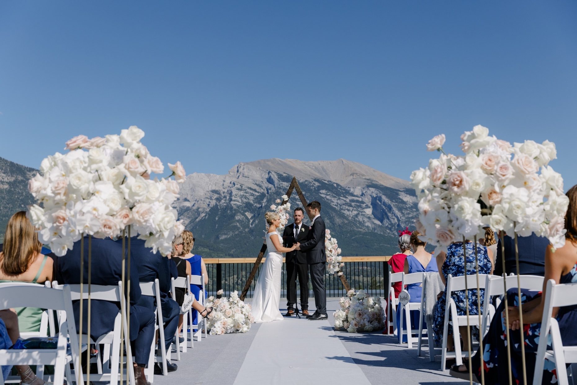 Wedding Vows Couple exchanging vows under a triangle floral arch at Lake Louise, with tall aisle arrangements of white and blue roses in the foreground.
