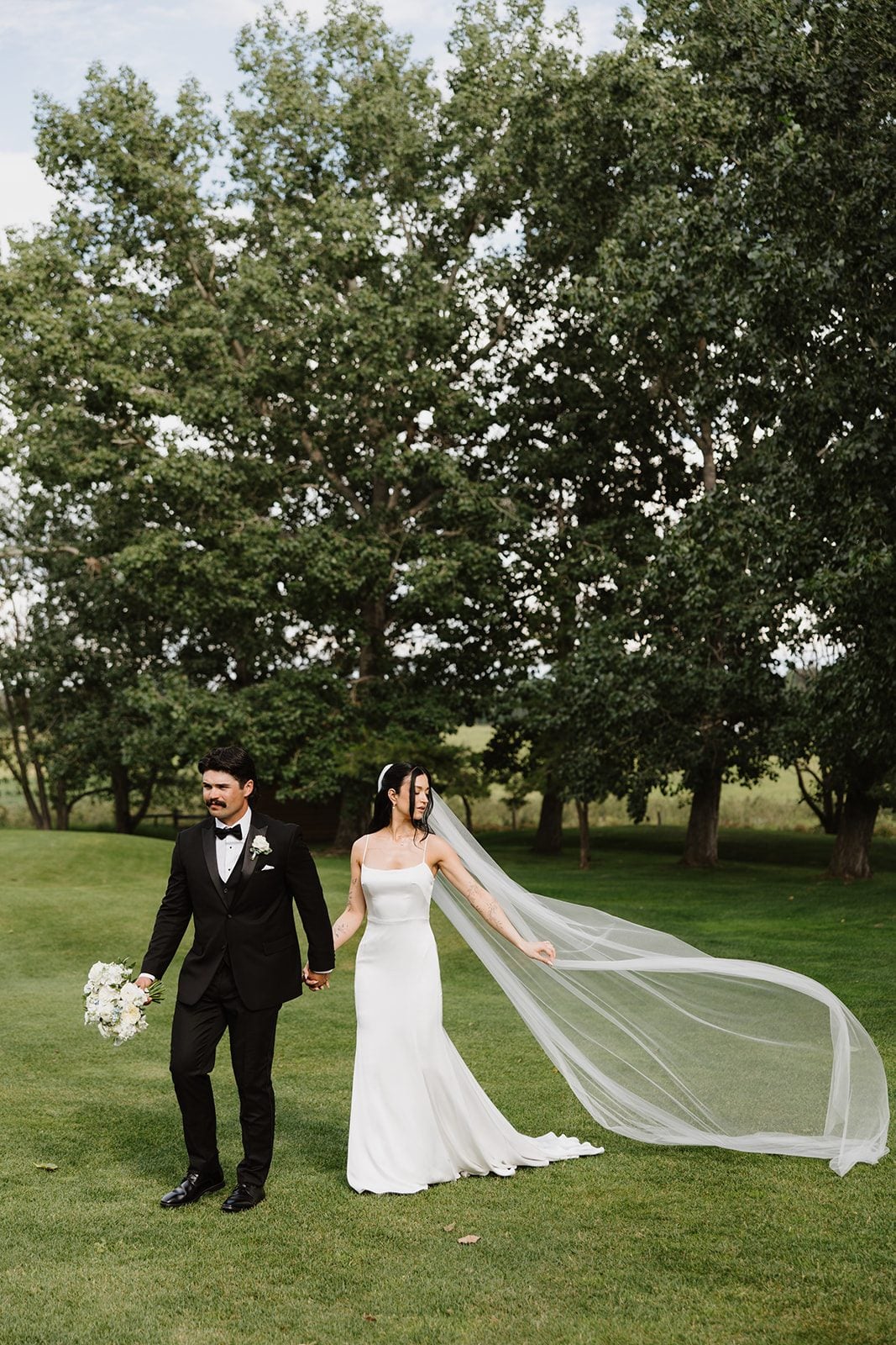 Cassidy + Adon Portrait of a white and green wedding at Hilltop. Bride and groom are walking across the grounds hand in hand, her veil billowing out behind her and he carries her white bridal bouquet.