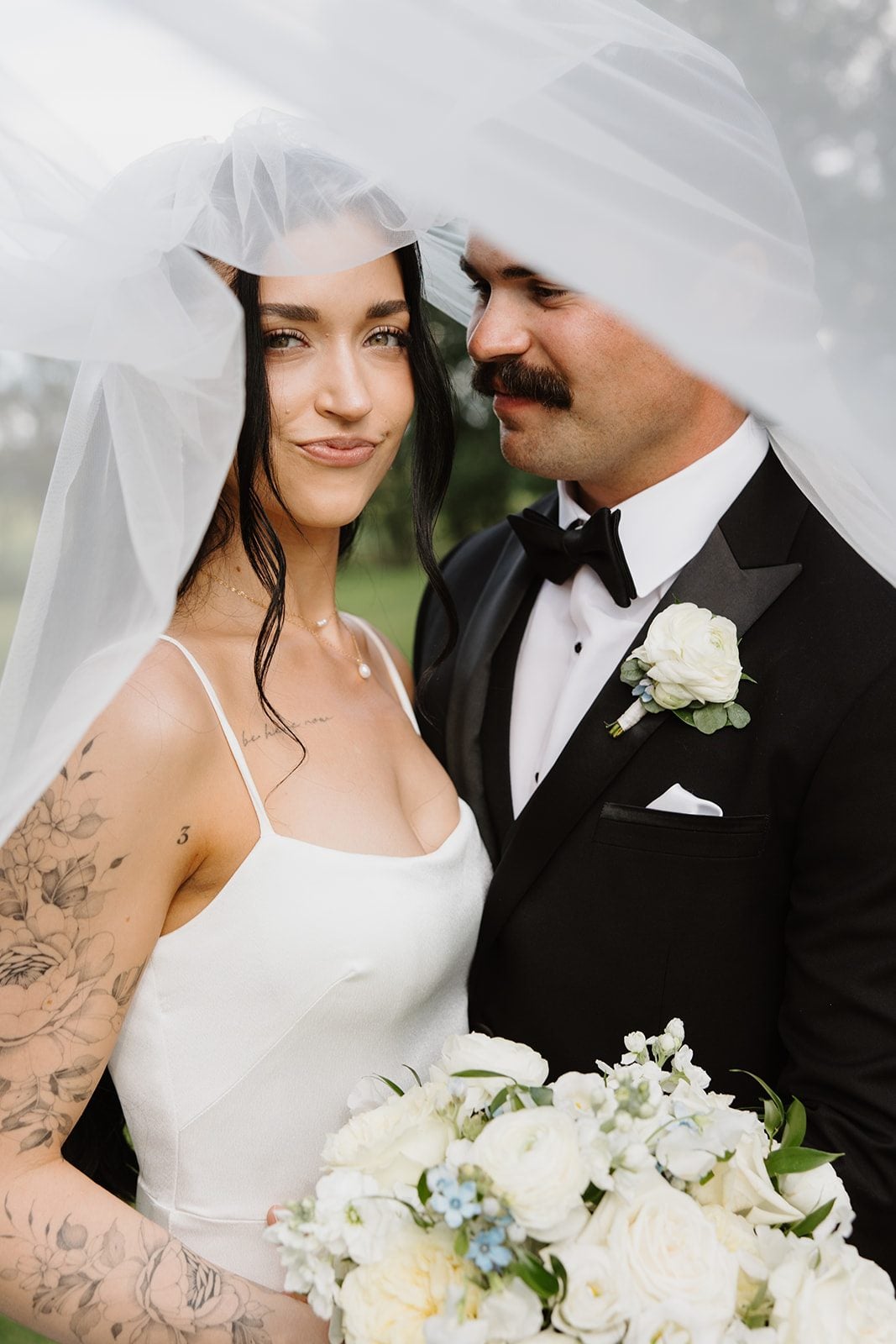 Cassidy + Adon Bride (holding a bridal bouquet of white flowers and lush greens) and groom (wearing black suit with white boutonnière) are close together with her veil framing around them.
