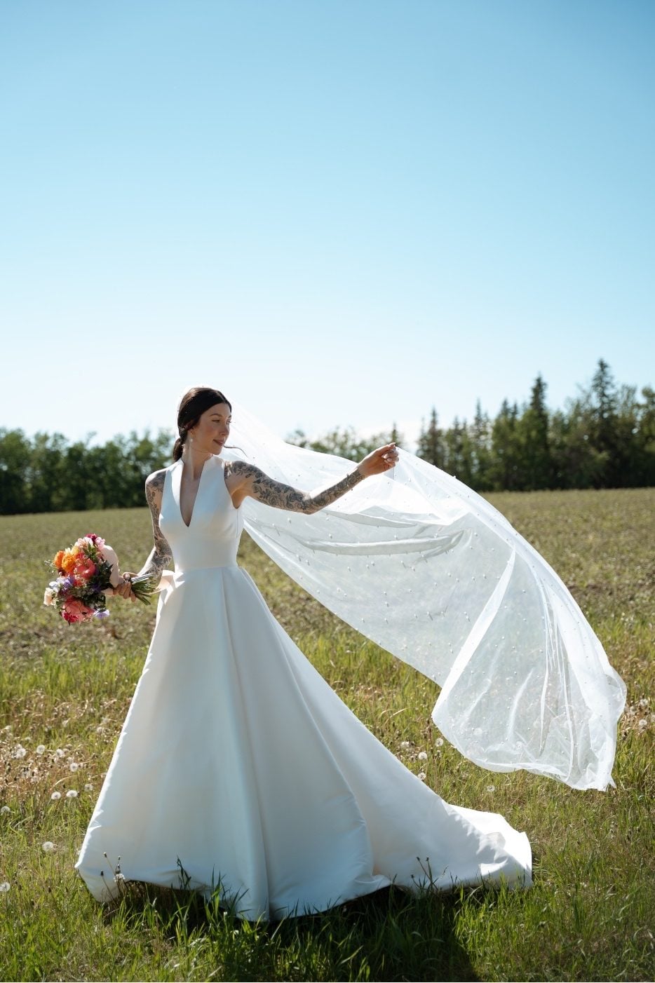 Bride with Colourful Bouquet Bride stands in the field at River's Edge, her colourful bouquet in one hand and her other hand holding her veil blowing in the wind behind her.
