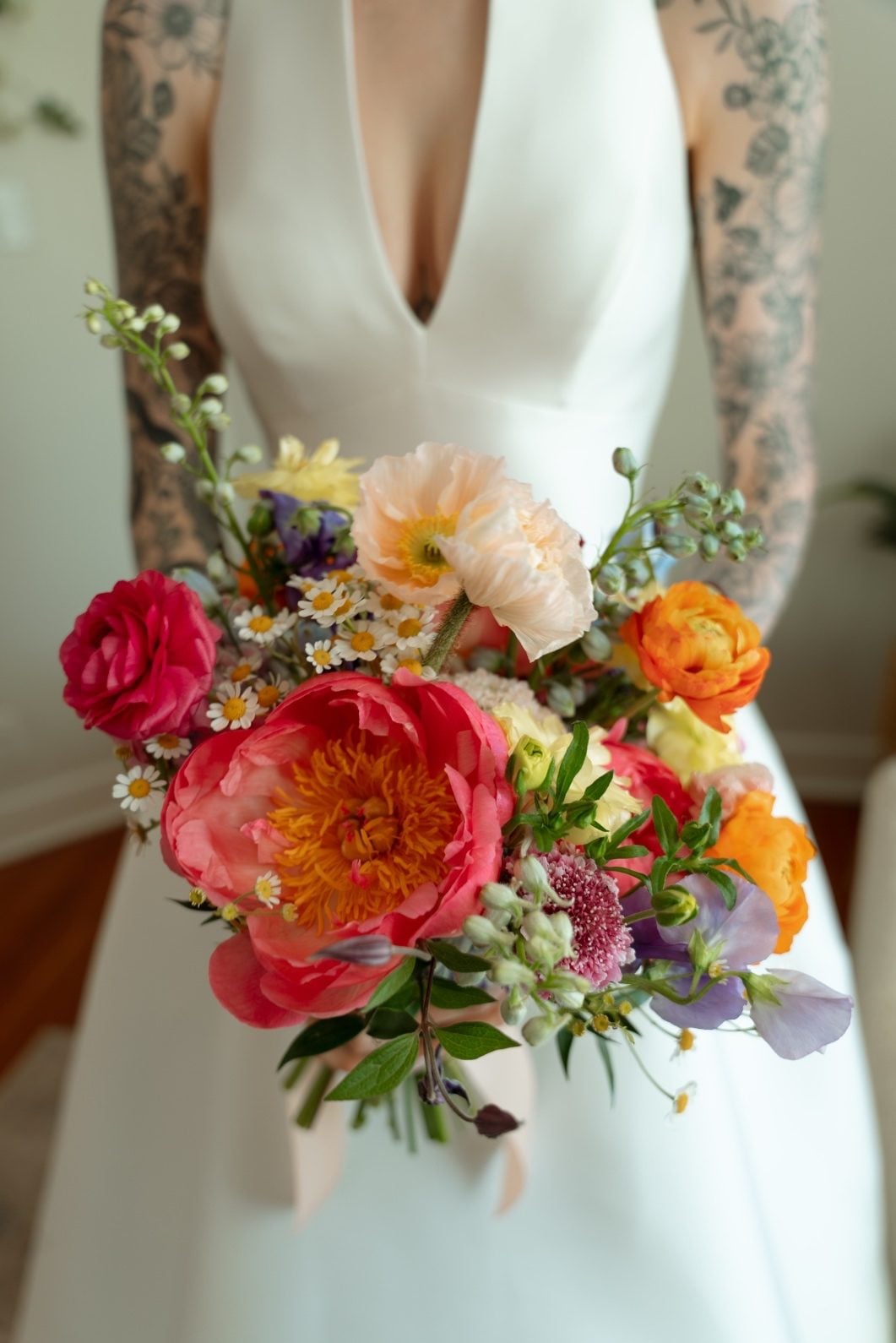 Colourful Bridal Bouquet with Coral Charm Peonies A close up of the brides stunning, colourful bouquet featuring a huge coral charm peony, ranunculus, poppies, spray roses, sweet peas, delphinium, and chamomile