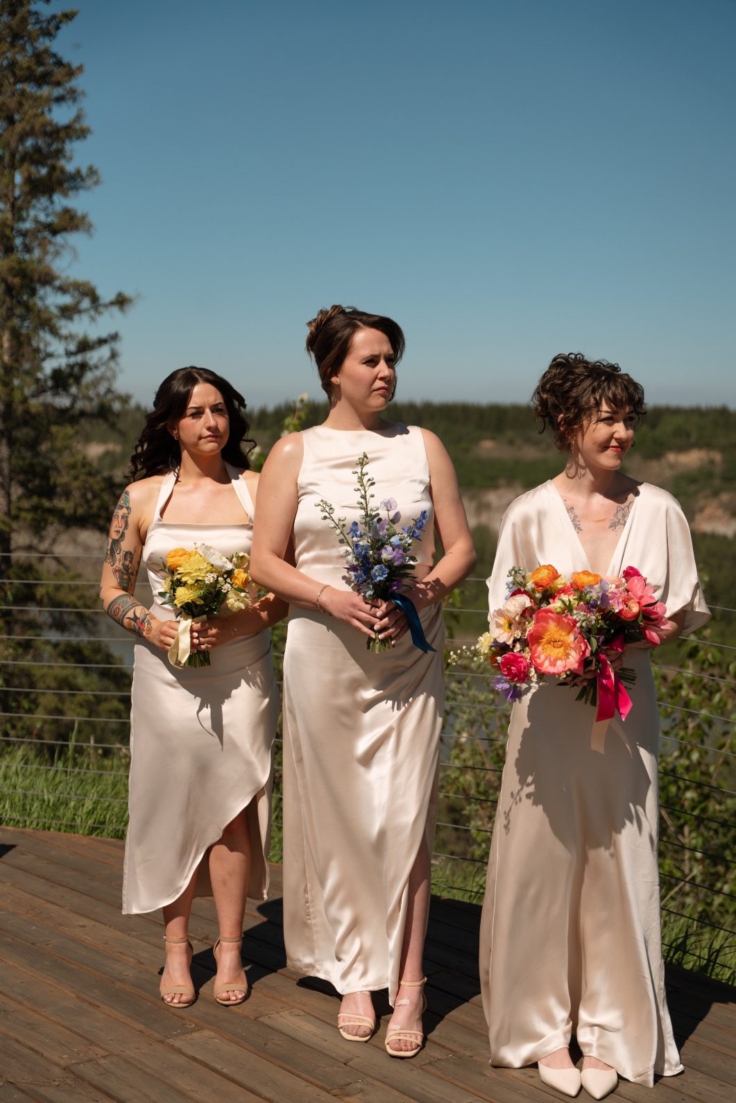 Colourful Bridesmaids The bridesmaids standing in a row during the ceremony in their champagne gowns. Each one carries a monochromatic, colourful bouquet - one yellow, one blue, one pink.