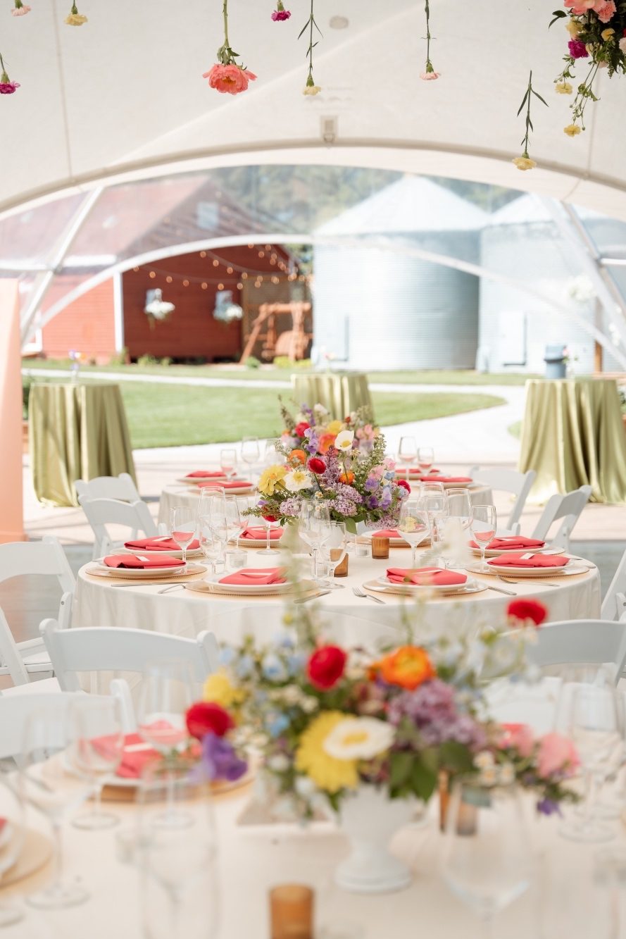 Colourful Centrepieces at River’s Edge Round tables with colourful, gardeny centrepieces, plates with coral folded napkins and white textured linens.