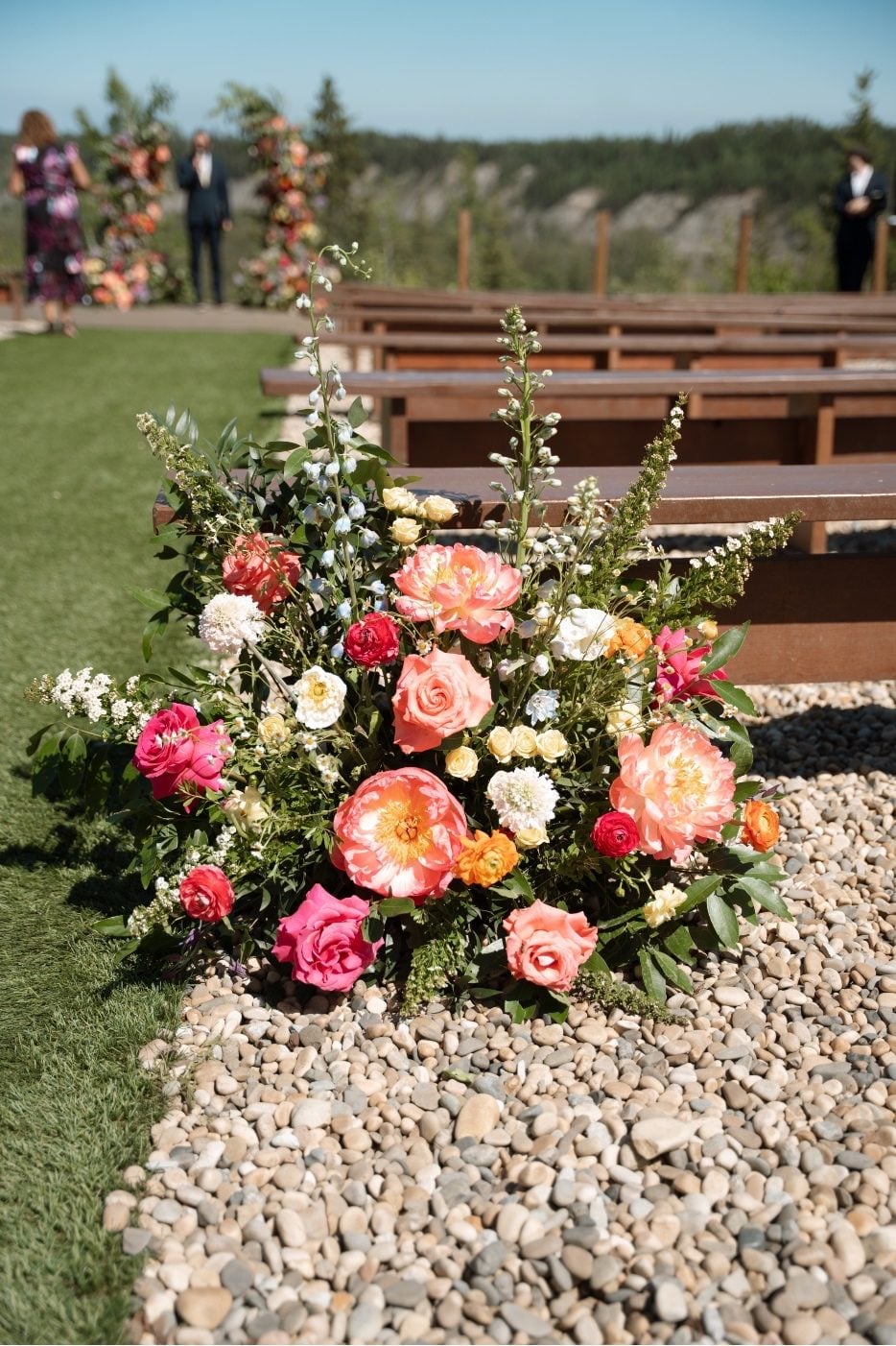 Colourful Ceremony Flowers A stunning arrangement at the start of the aisle at River's Edge featuring coral peonies, delphinium, ranunculus, roses, scabiosa, chamomile, and flowering spirea.