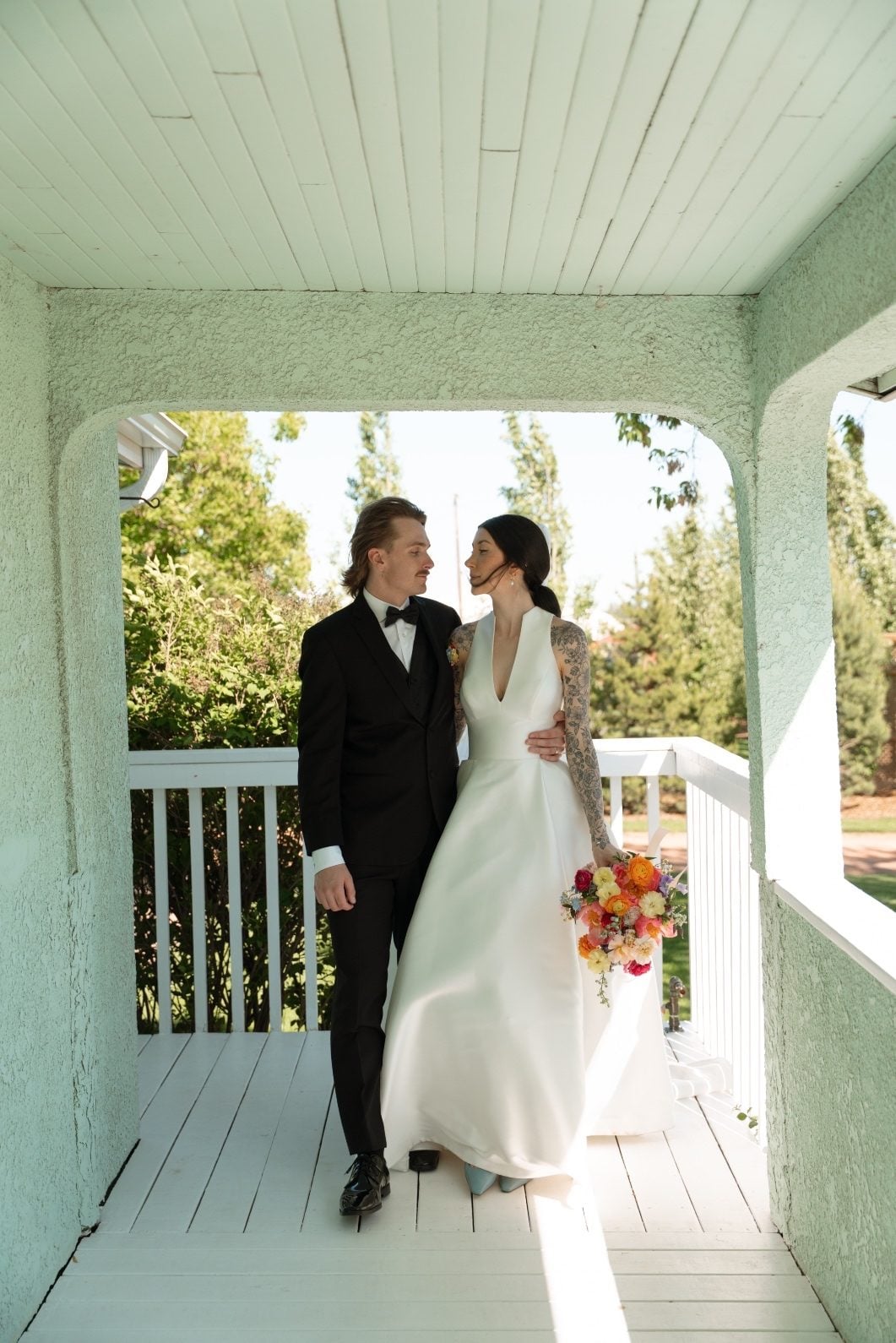 Colourful Wedding at River’s Edge The bride and groom standing on a covered porch, the bride holds a colourful garden inspired bouquet.