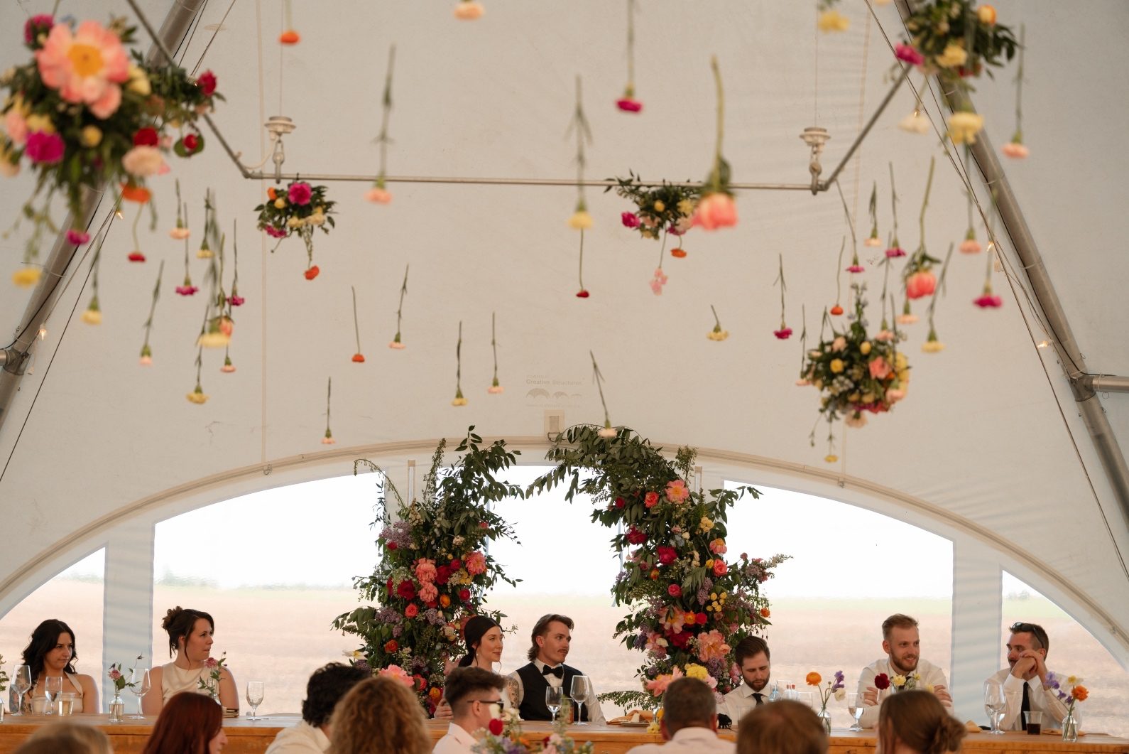 Suspended Flowers at Colourful Reception The bride and groom sit at the head table, with the stunning, colourful broken arch framing them from behind, and flowers suspended from the tents ceiling.