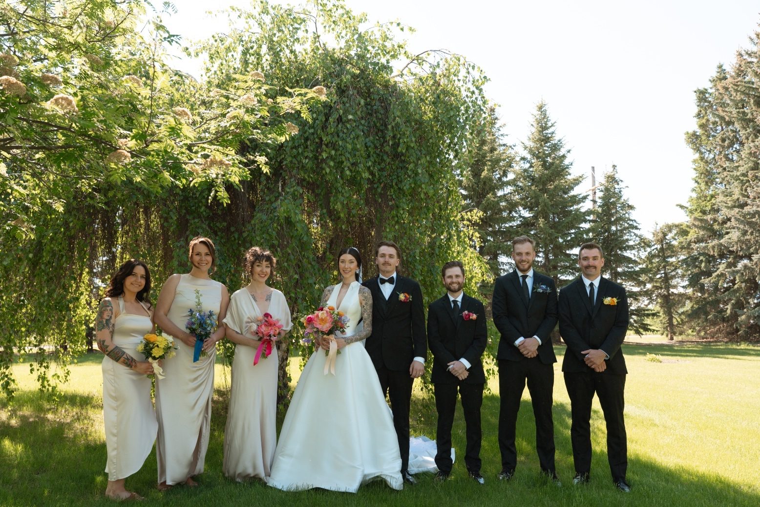 Wedding Party at River’s Edge The bride and groom stand under a large tree, with their wading party on either side of them, holding colourful bouquets and wearing colourful boutonnières.