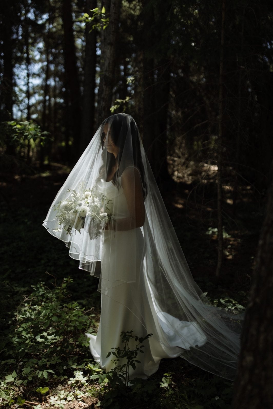 Veiled Green and White Bride Bride stands in the forest on the grounds at Archway Manor with her veil over her face and bouquet.