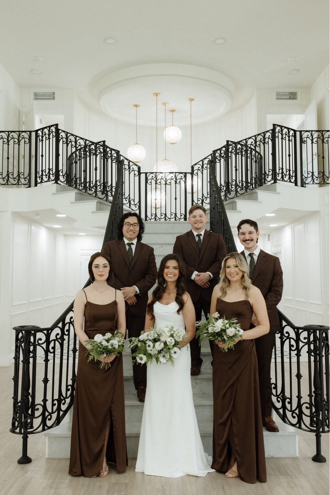 White and Green Bridal Party at Archway Manor A bride stands with her bridal party, wearing chocolate brown attire, on the grand staircase at Archway Manor.
