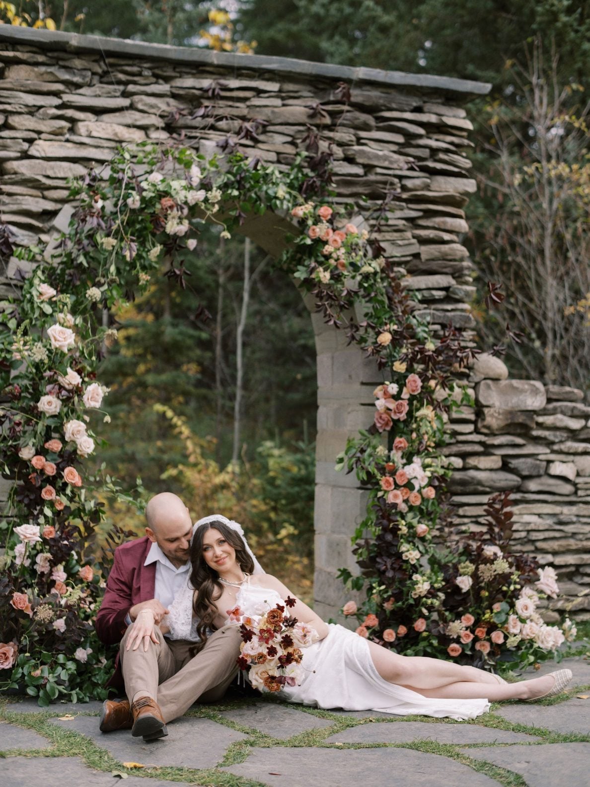 Justine Milton Marsala Couple at The Valley Marsala true to hue photoshoot at The Valley Weddings, couple sits in front of stone arch adorned in blush and burgundy flowers.