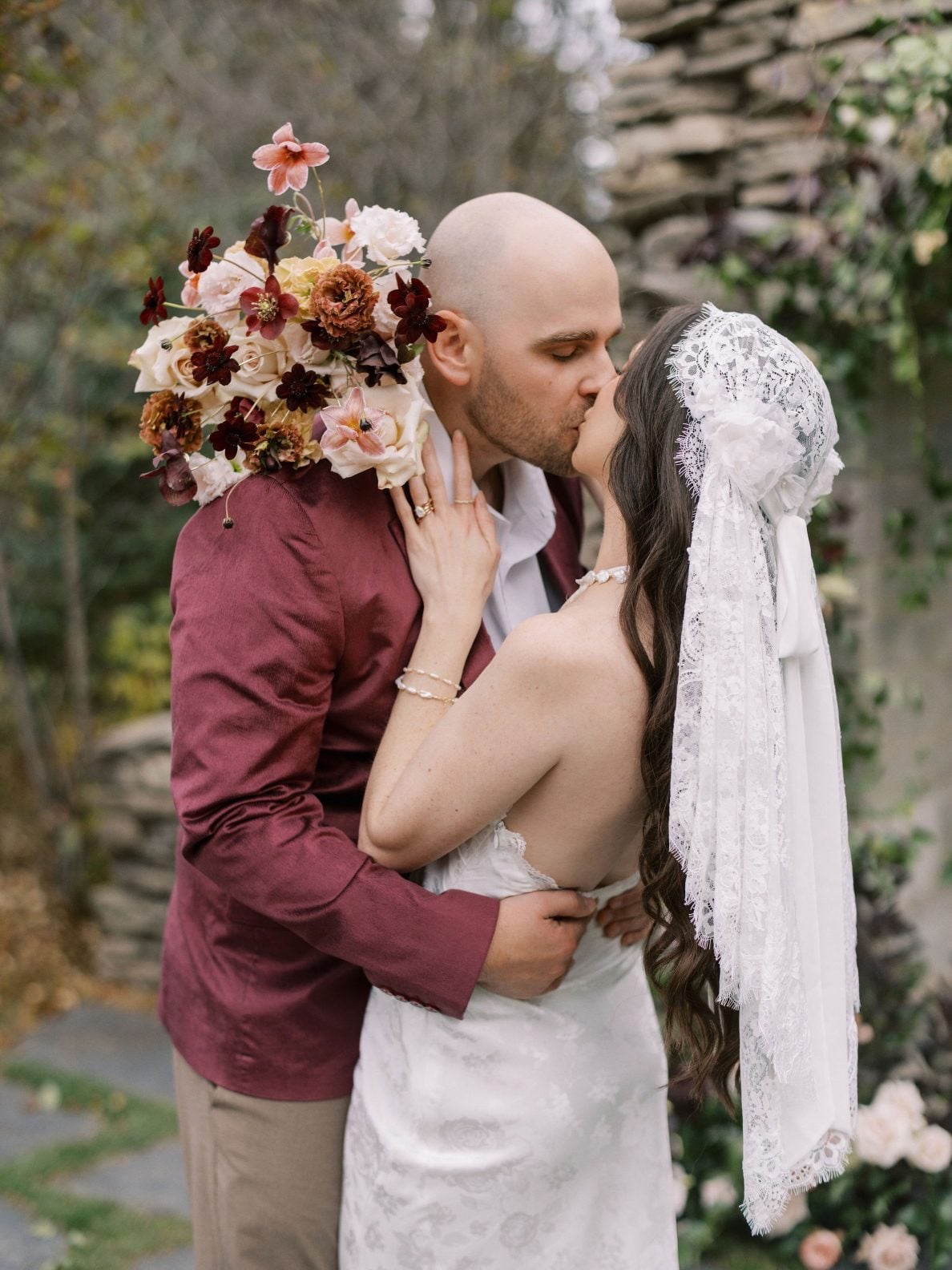 Justine Milton Marsala Couple Model groom kisses bride in her Juliet veil while she holds her blush and burgundy bouquet.