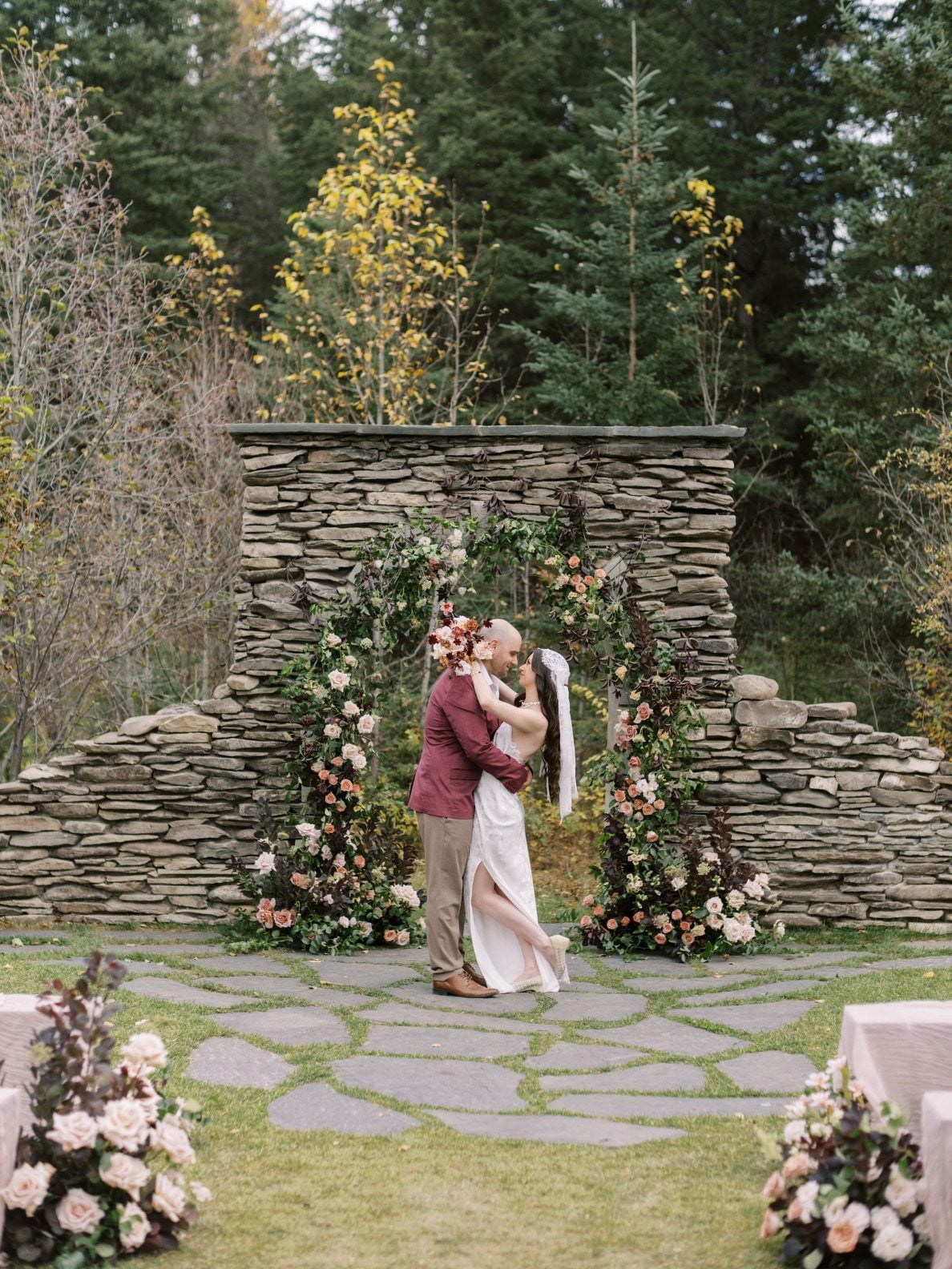 Justine Milton Marsala True to Hue Couple at Stone Arch Model bride and groom share a kiss in front of the stone arch decorated in flowers for the ceremony portion of the True to Hue photoshoot.