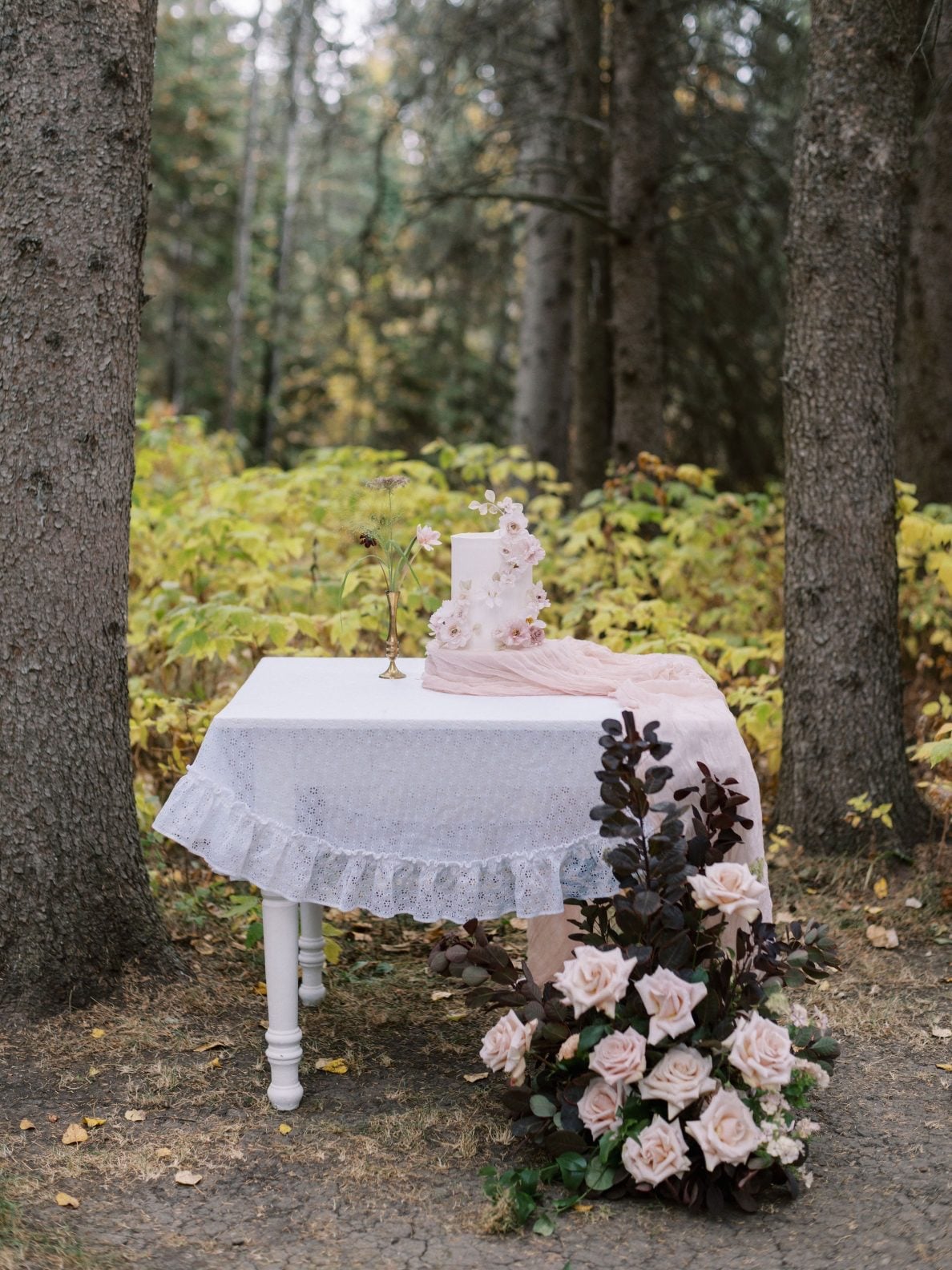 Justine Milton True to Hue Cake Table A simple table with lace table cloth, tall arrangement on the ground next to the table in beige, blush and burgundy, simple bud vase on the table, and stunning two layer wedding cake accented by handmade flowers.