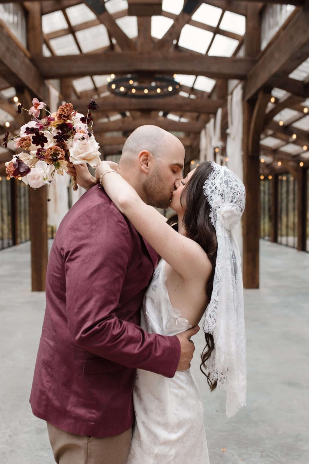 Nikki Collette Marsala True to Hue Couple at The Valley Bride holds her beige, blush and burgundy bridal bouquet of chocolate cosmos, roses, tulips and lisianthus, while sharing a kiss with the groom in front of the reception space at The Valley.