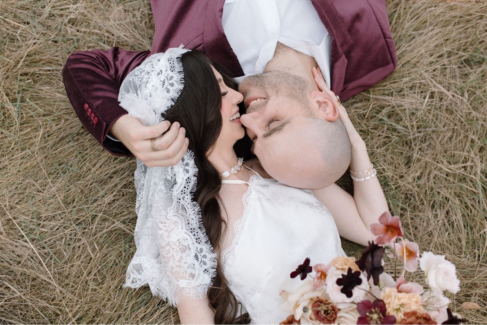 Nikki Collette True to Hue Marsala Couple Portraits Bride holds her blush and burgundy bridal bouquet as her and the groom lay in the grass smiling as they are about to kiss.