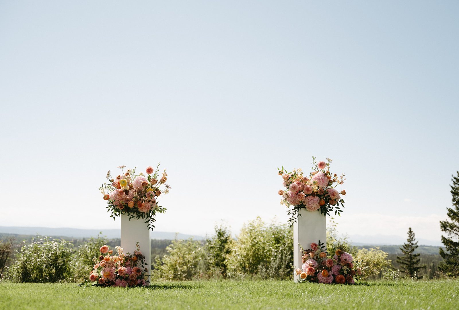 Big, gardeny pink and peach arrangements both on top and at the base of white pillars, with clear blue skies behind them.