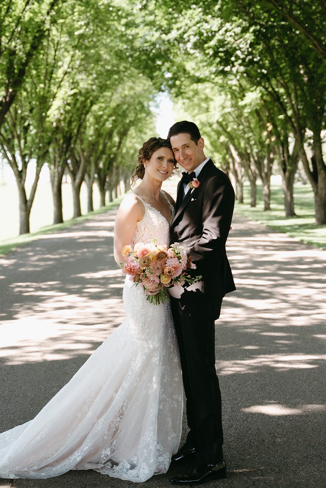 Bride and groom stand closely on a path lined with trees smiling. She holds a pink and peach garden inspired bridal bouquet.