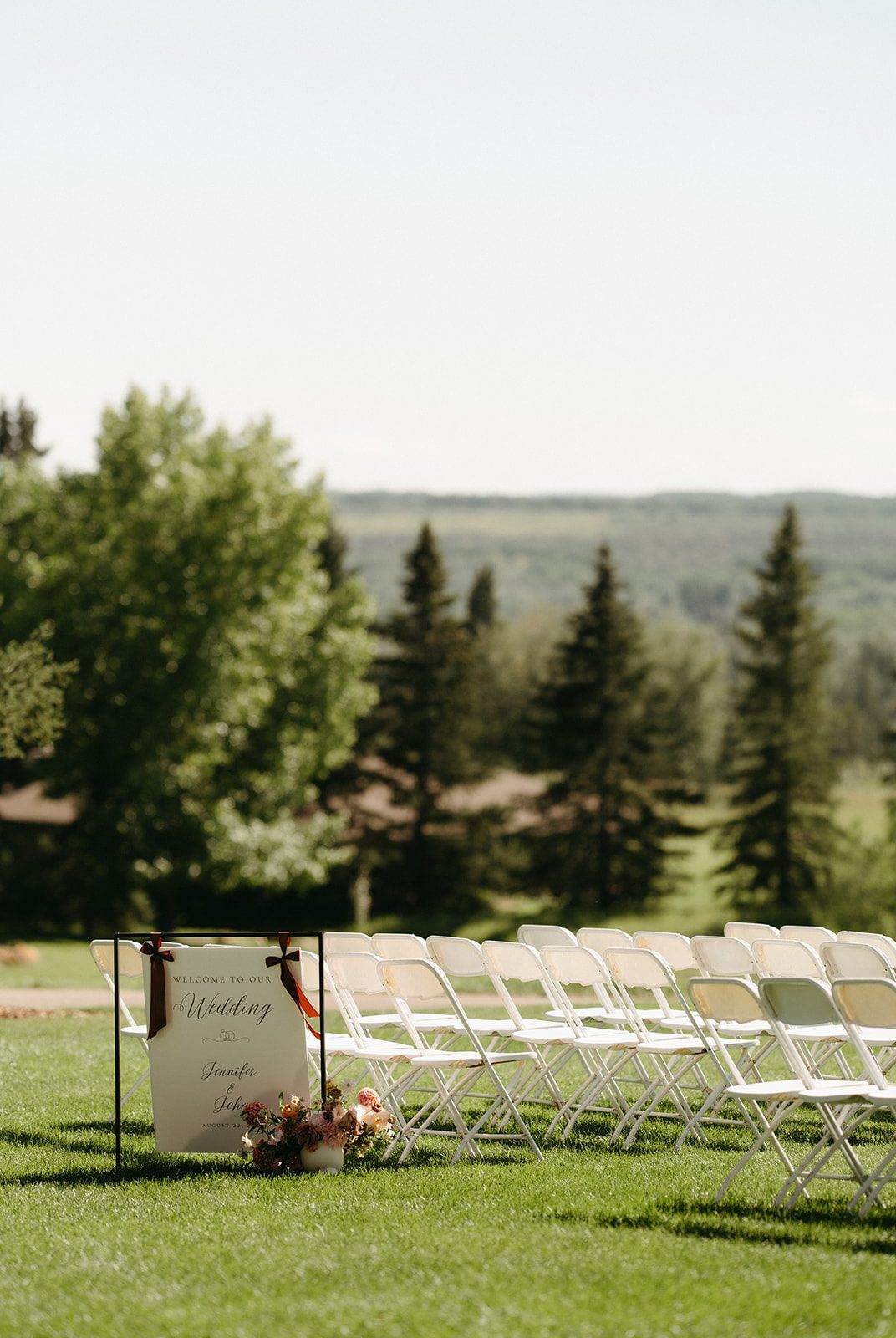 Outdoor ceremony at Pinebrook Golf & Country Club, a welcome sign with a pink and peach floral arrangement at its base in front of neat rows of white chairs.