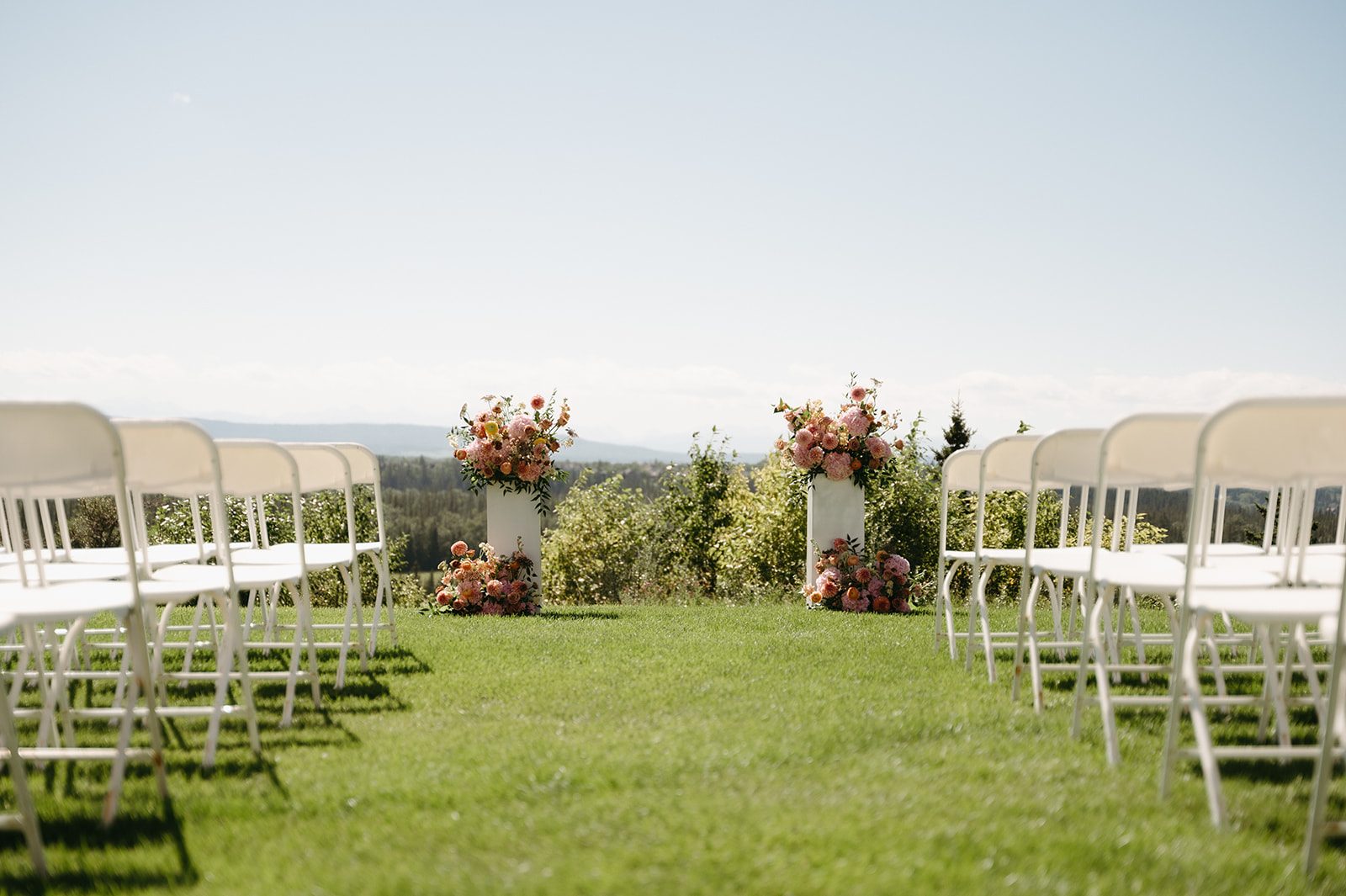 Looking down the aisle at big, beautiful pink and peach arrangements on top of white pillars, with arrangements also in the grass down at their base.