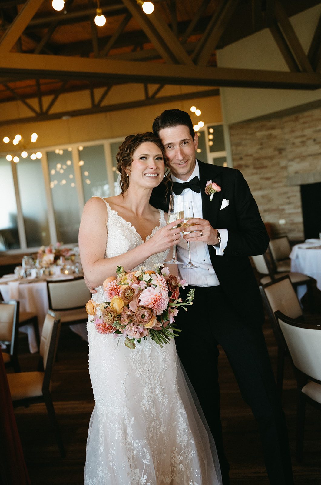 Bride holds her pink and peach bridal bouquet in one hand while she clinks champagne glasses with the groom with her other.
