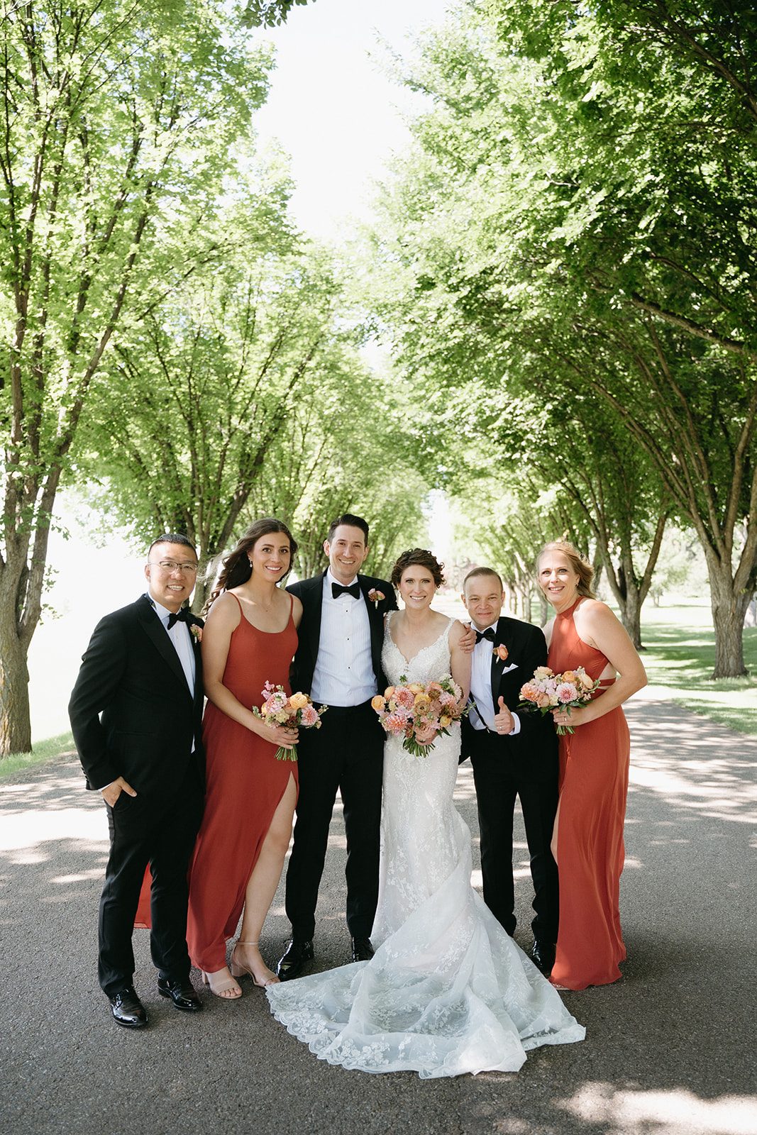 Bride and groom stand with their wedding party - the groomsmen wearing black tuxes and the bridesmaids wearing full length terracotta gowns.
