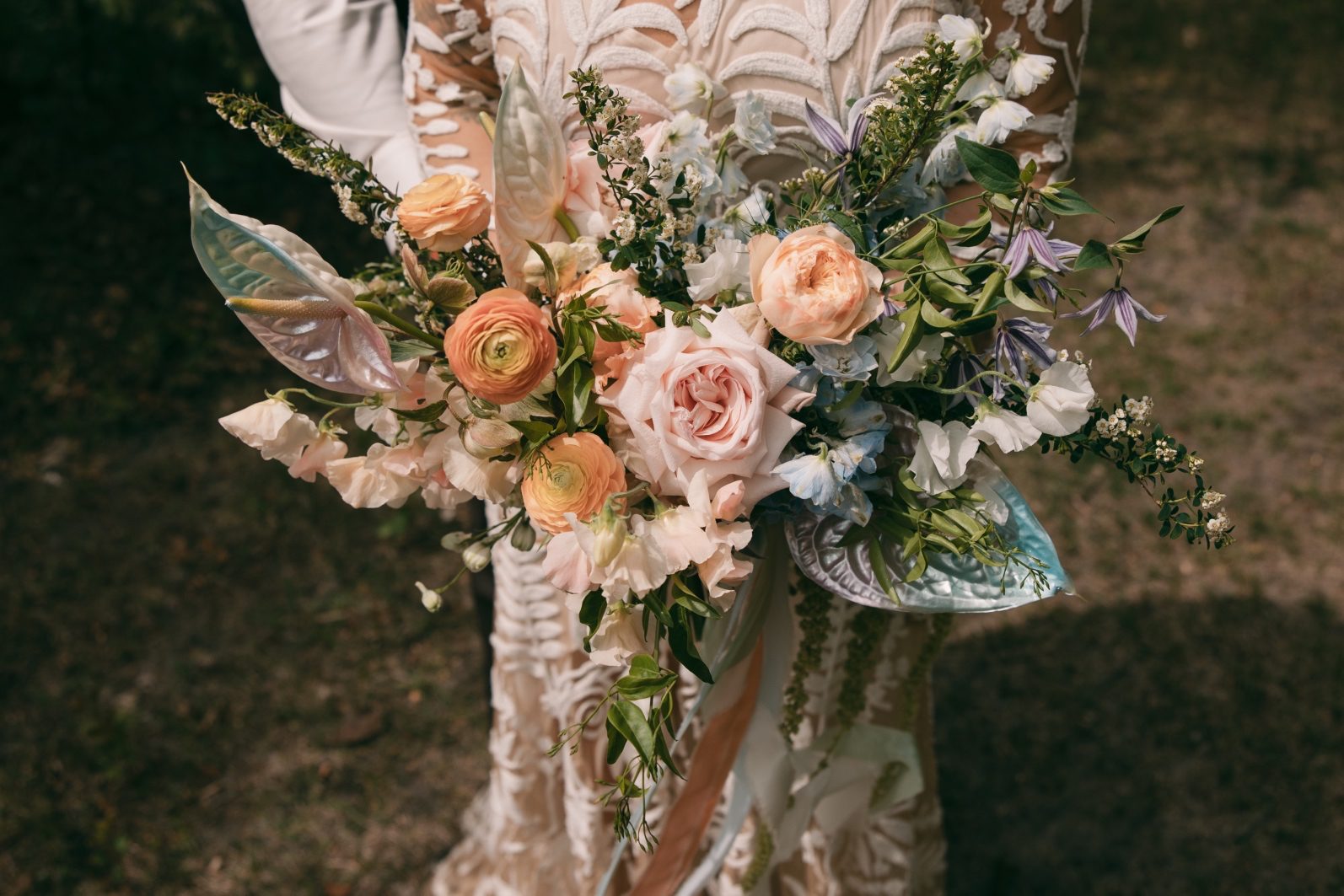Rainbow ombre bridal bouquet with peach ranunculus, blush roses, blue delphinium, clematis, spirea, sweet peas and iridescent anthurium.