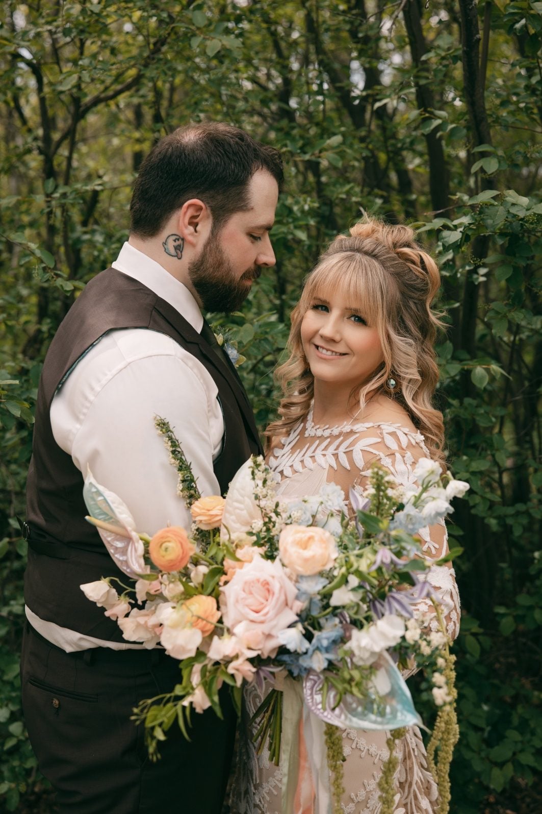 Leigha and Tyler standing close together while she smiles at the camera holding a rainbow ombre bridal bouquet of anthurium, spirea, roses, ranunculus, sweet peas, and clematis.