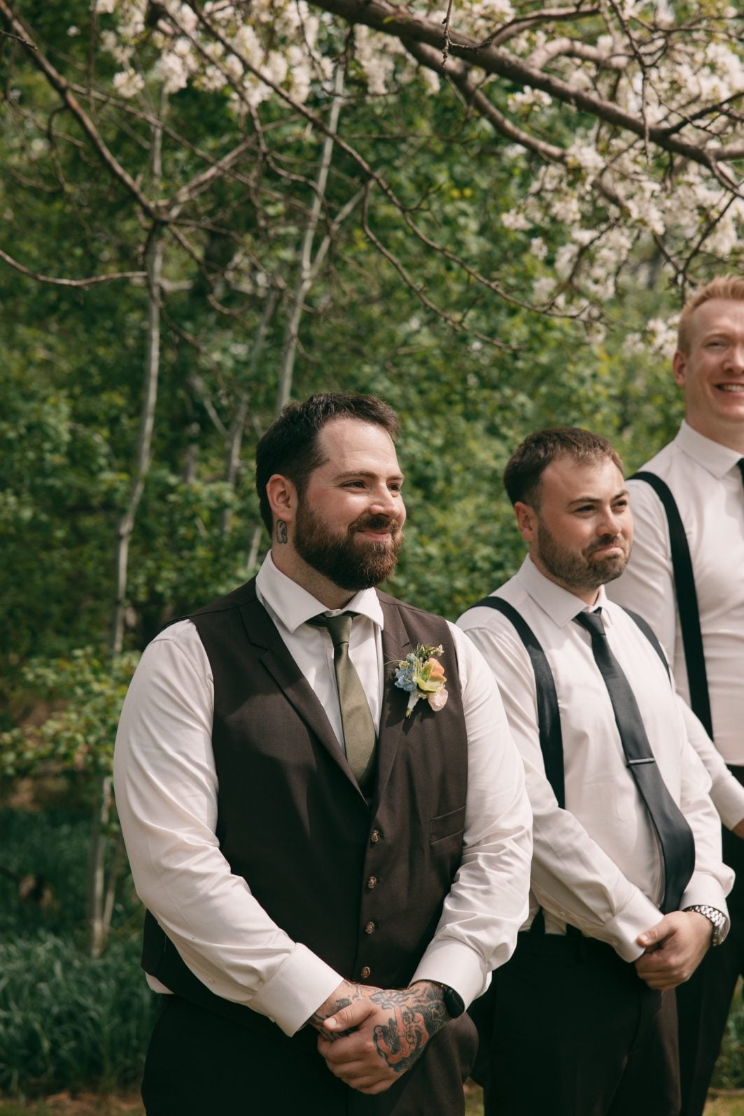 Groom smiling besides his groomsmen in relaxed wedding attire with suspenders and a pastel floral boutonnière as he watched his bride coming down the aisle.
