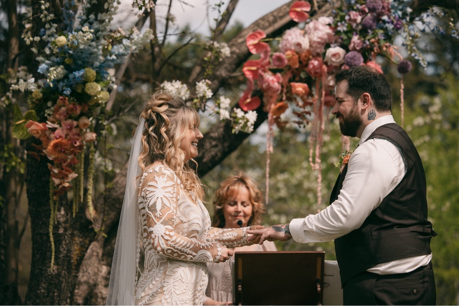 Bride and groom exchanging rings beneath a colourful floral installation during their intimate outdoor wedding ceremony.