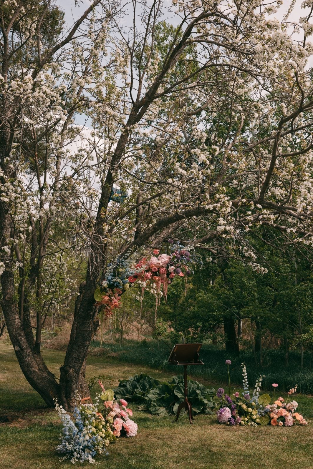 Colourful outdoor wedding ceremony beneath a flowering tree with pastel rainbow floral installations framing the ceremony space.