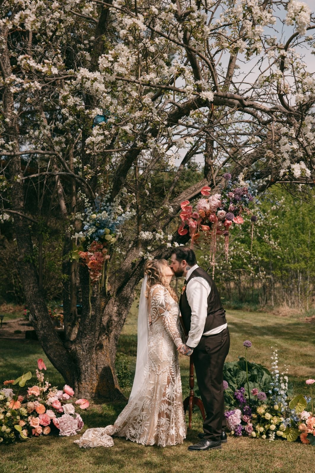 Newlyweds sharing their first kiss beneath a flowering tree framed by colourful spring wedding flowers.