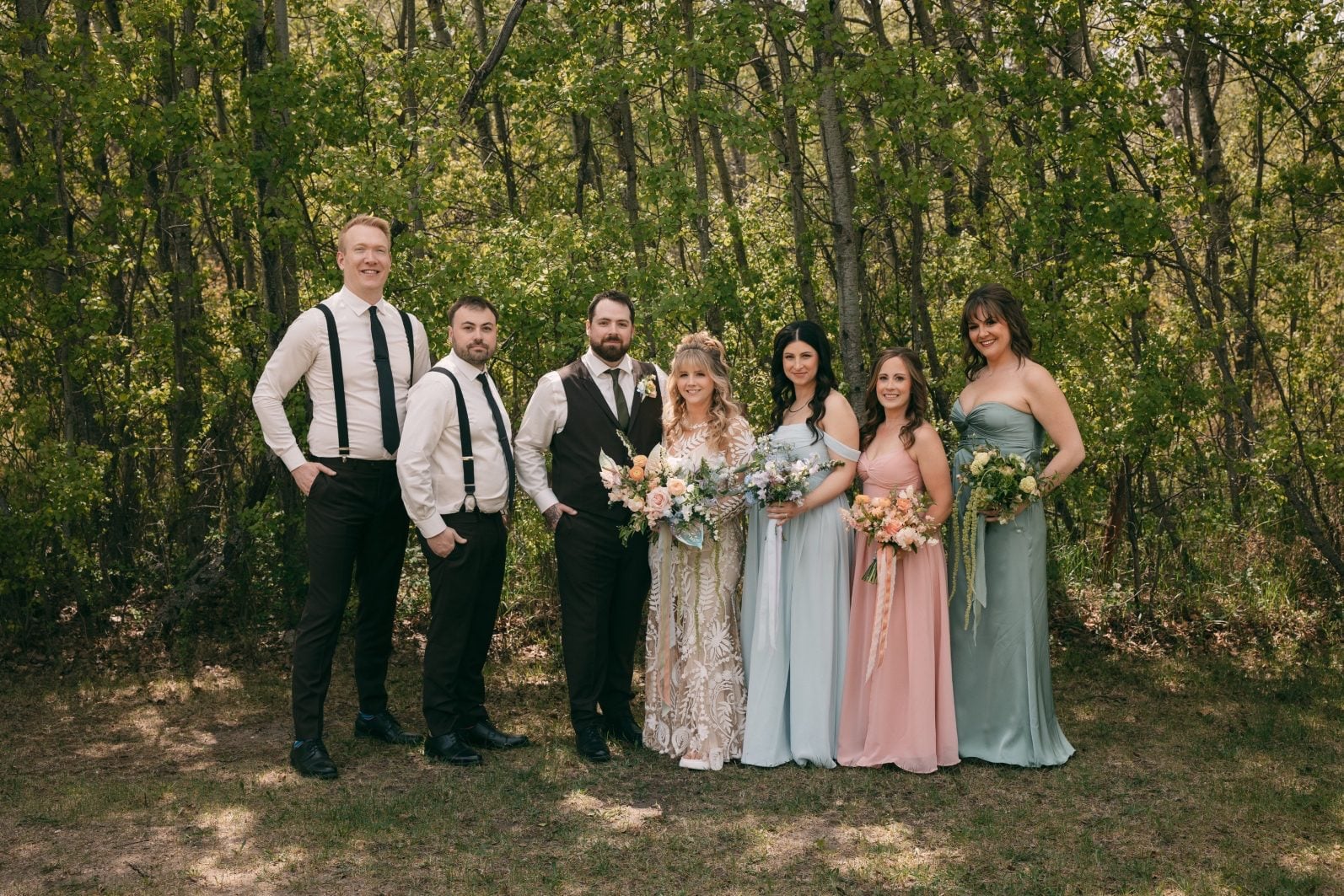Bride, Groom and wedding party surrounded by trees with coordinating pastel dresses and colourful bouquets.
