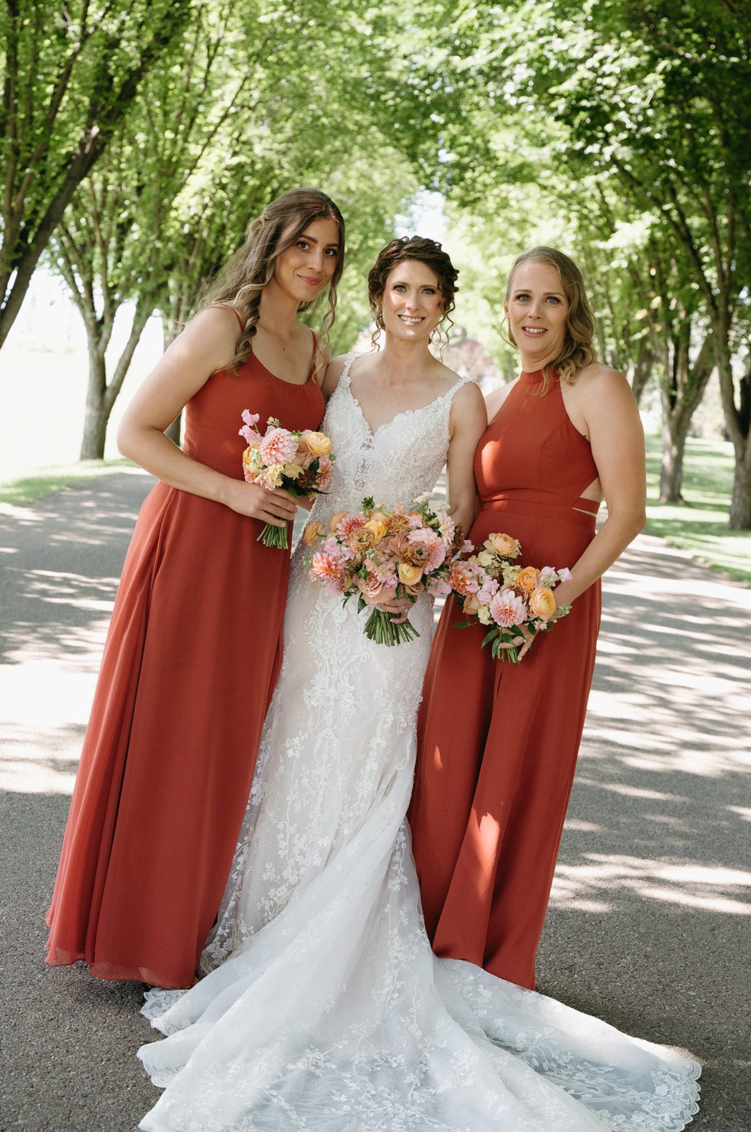 Bride stands with her beautiful bridesmaids wearing full length terracotta gowns, all holding pink and peach garden inspired bouquets.