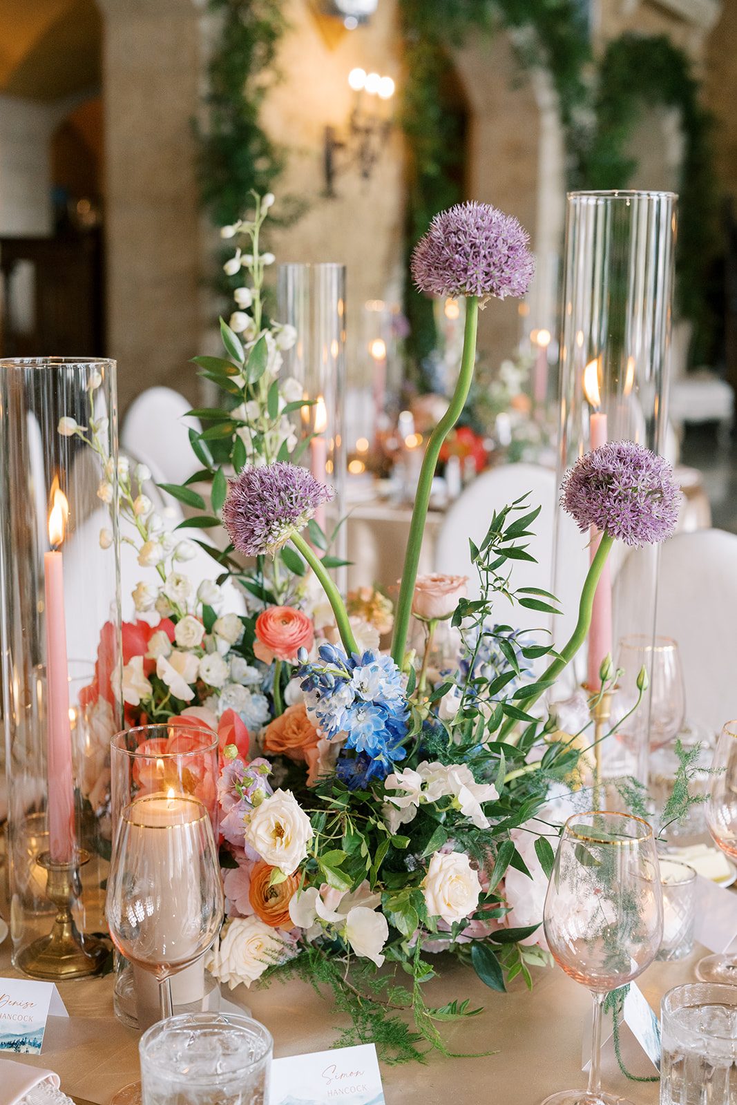 Lottie Garden inspired wedding centrepiece with purple allium, blue delphinium, coral and blush flowers, greenery and pink taper candles.