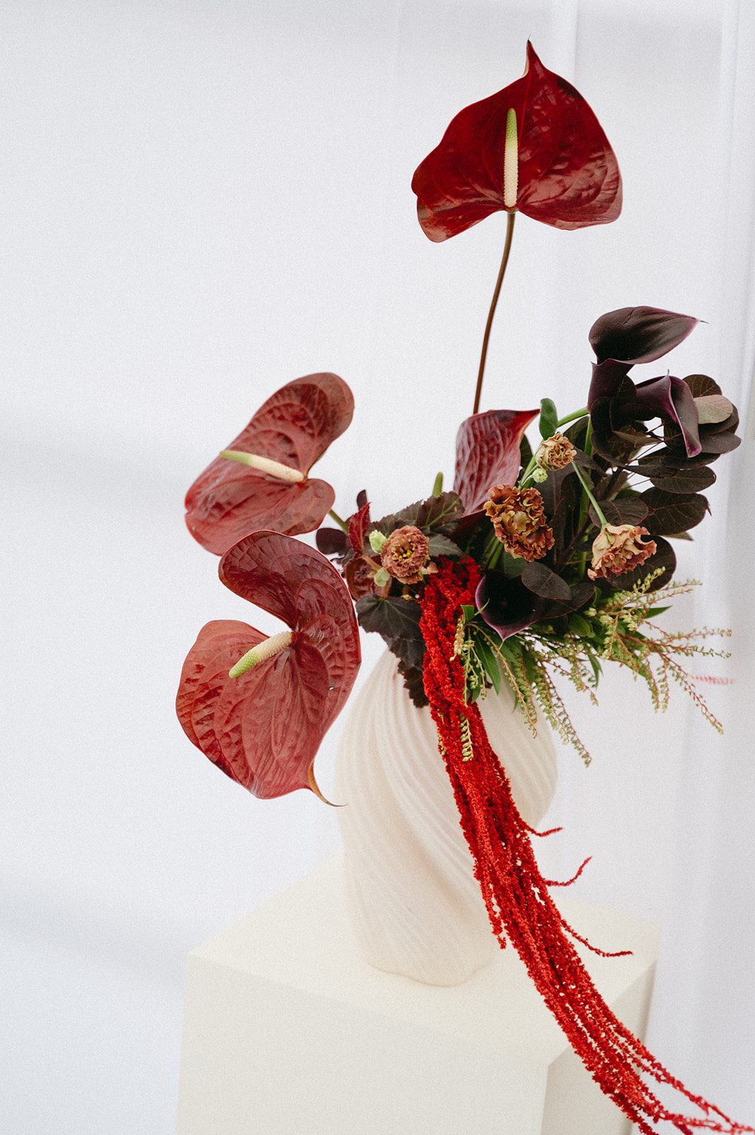 Andi + Logan Sculptural wedding flower arrangement featuring burgundy anthurium, calla lilies and cascading red amaranthus in a white ceramic vessel.