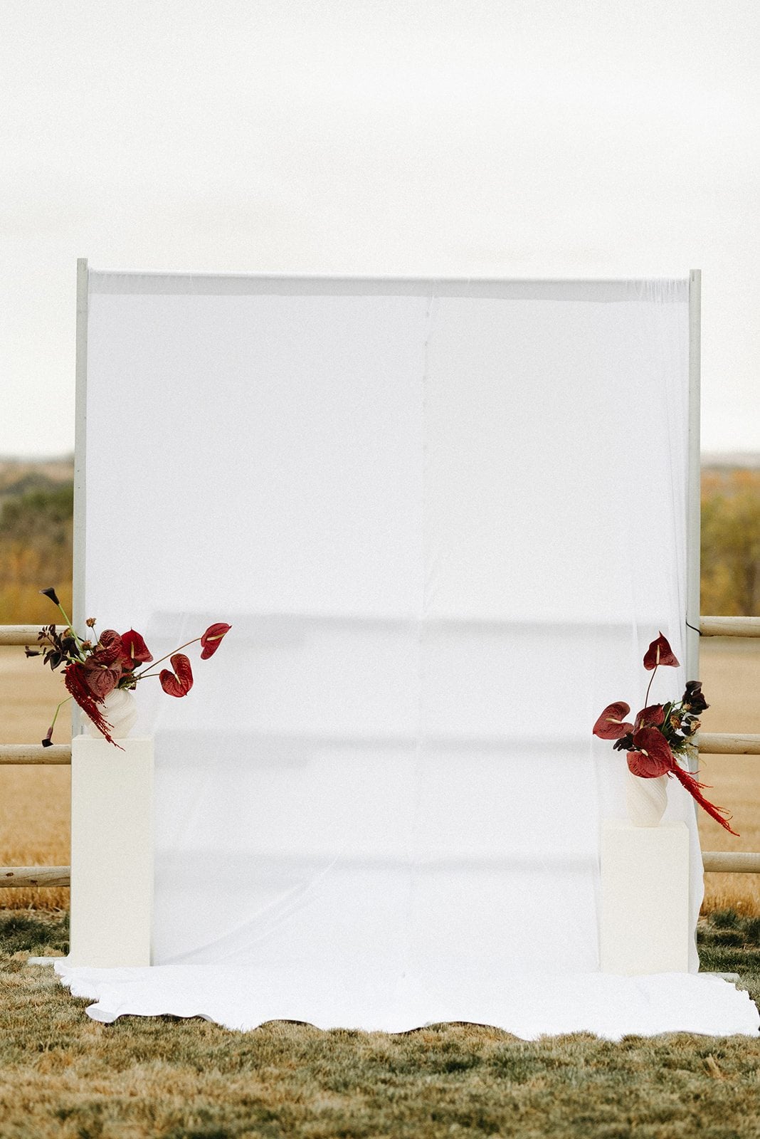 Andi + Logan Minimalist outdoor wedding backdrop framed by two asymmetrical burgundy sculptural floral arrangements on white pillars.