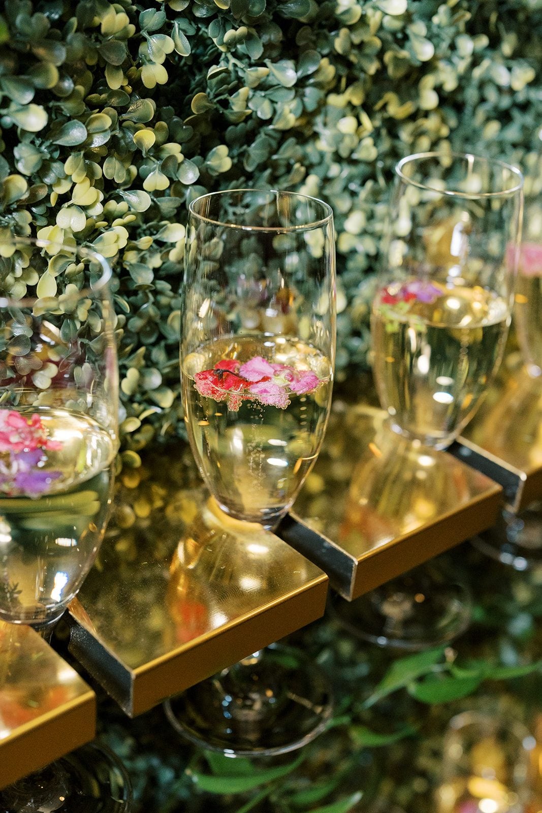 Lottie Champagne glasses garnished with colourful edible flowers on a greenery covered champagne wall.
