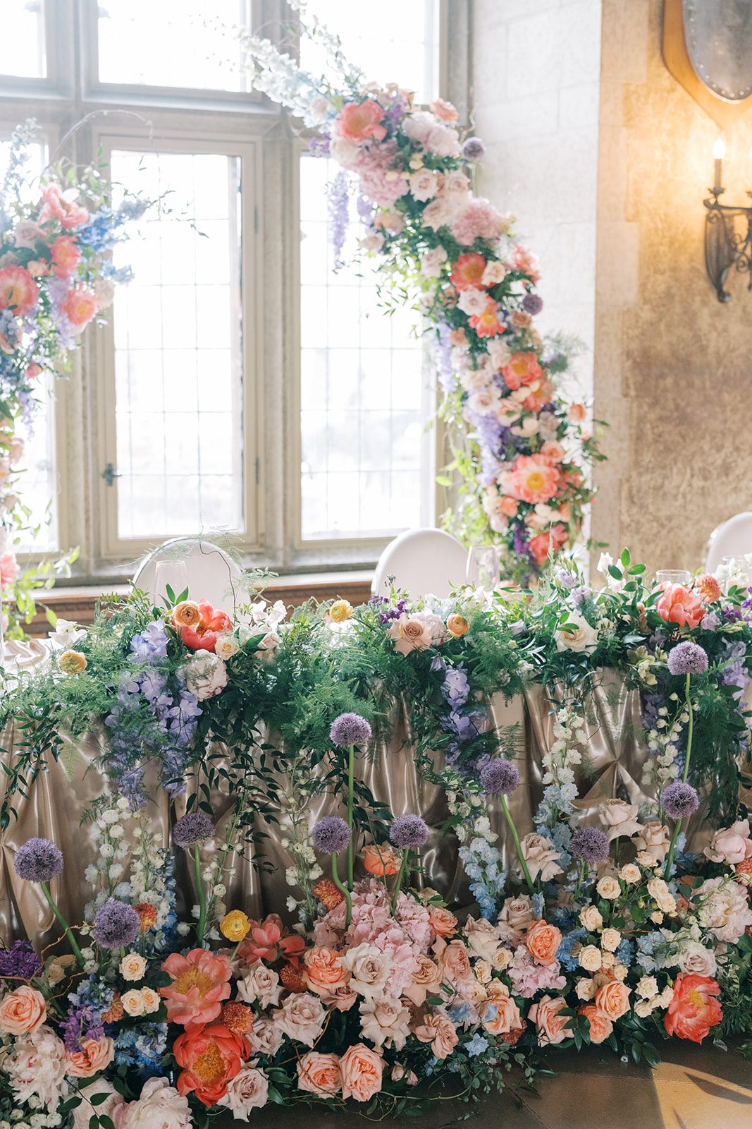 Lottie Colourful garden inspired wedding flowers cascading around the head table with coral peonies, roses, delphinium and purple allium.