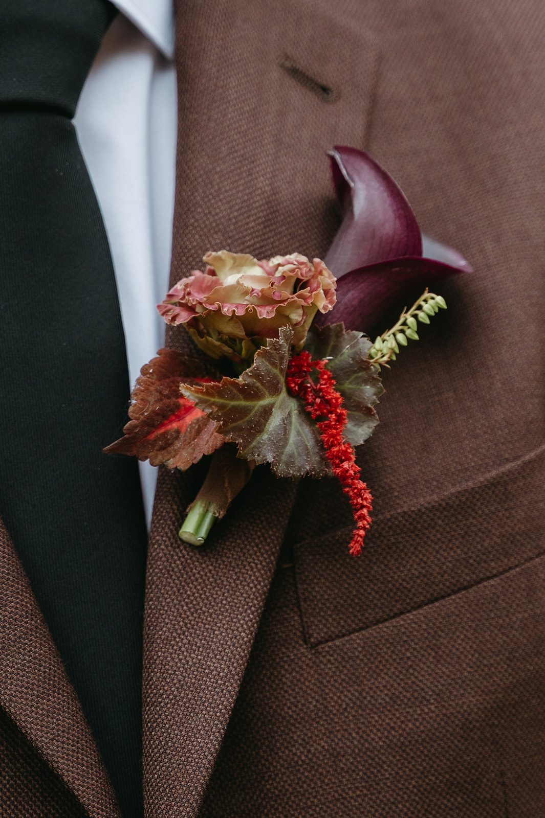 Andi + Logan Fall wedding boutonnière with burgundy calla lily, mocha lisianthus, begonia leaves and red amaranthus against the grooms brown suit.