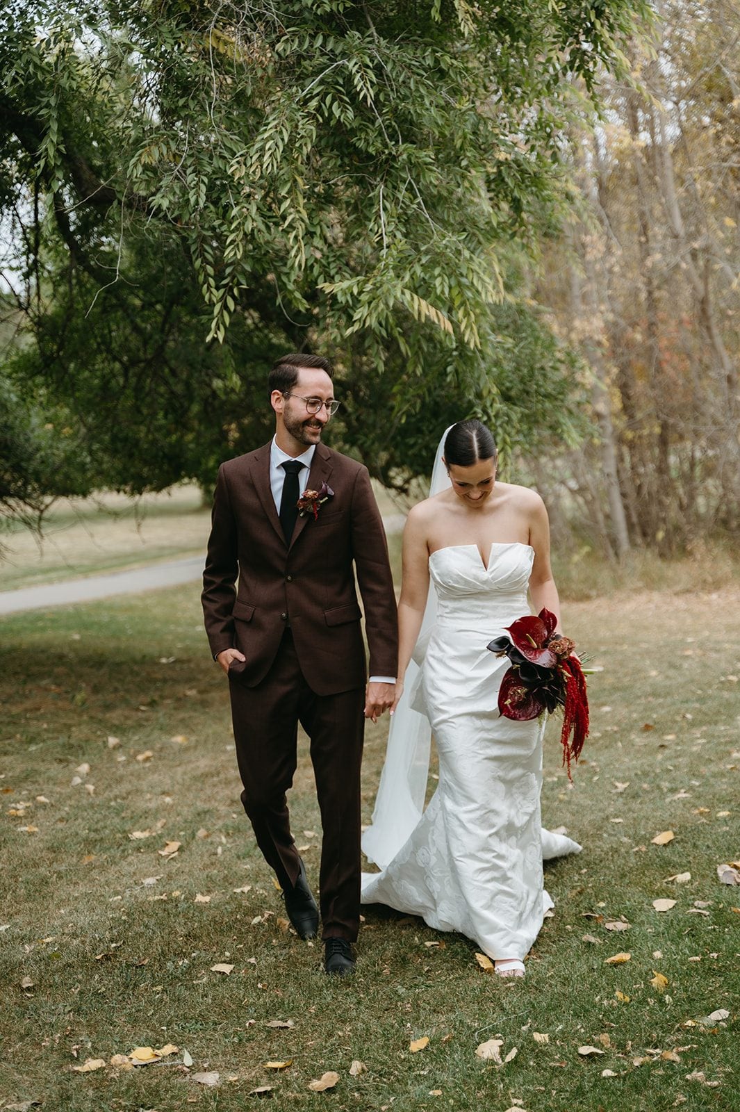 Andi + Logan Bride and groom walking together with a dramatic burgundy sculptural bridal bouquet during their fall wedding portraits.
