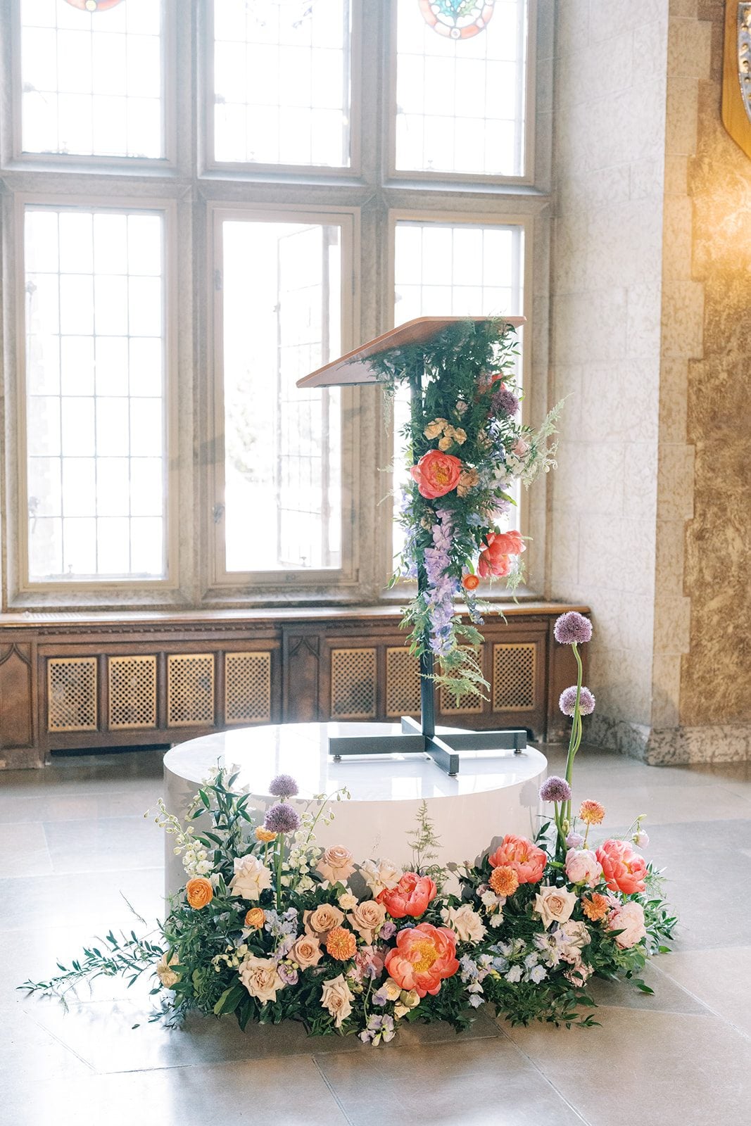 Lottie Garden inspired wedding flowers surrounding a podium with coral peonies, roses, purple allium, delphinium and greenery.