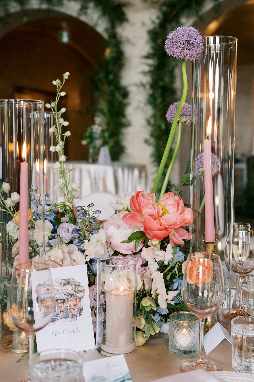 Lottie Colourful garden style wedding centrepiece with coral peonies, blue delphinium, blush flowers, alliums and pink taper candles.