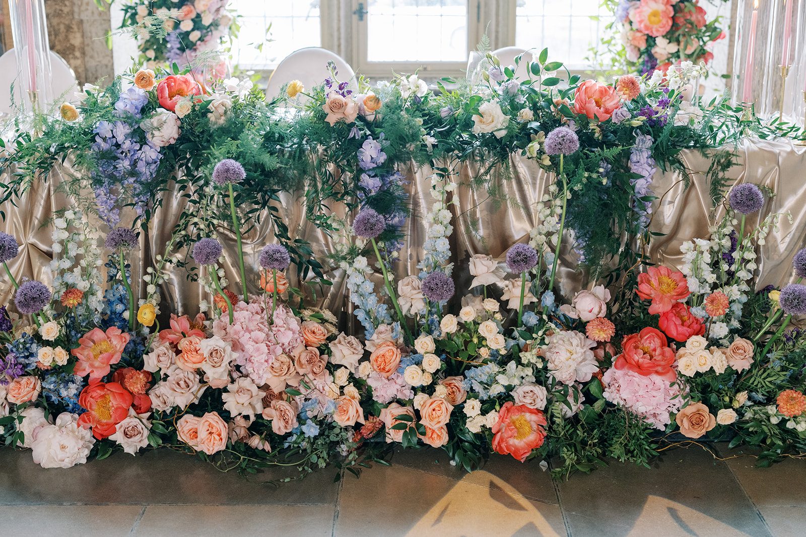 Lottie Abundant garden inspired head table flowers in coral, blush, lavender and blue with cascading greenery and tall alliums.
