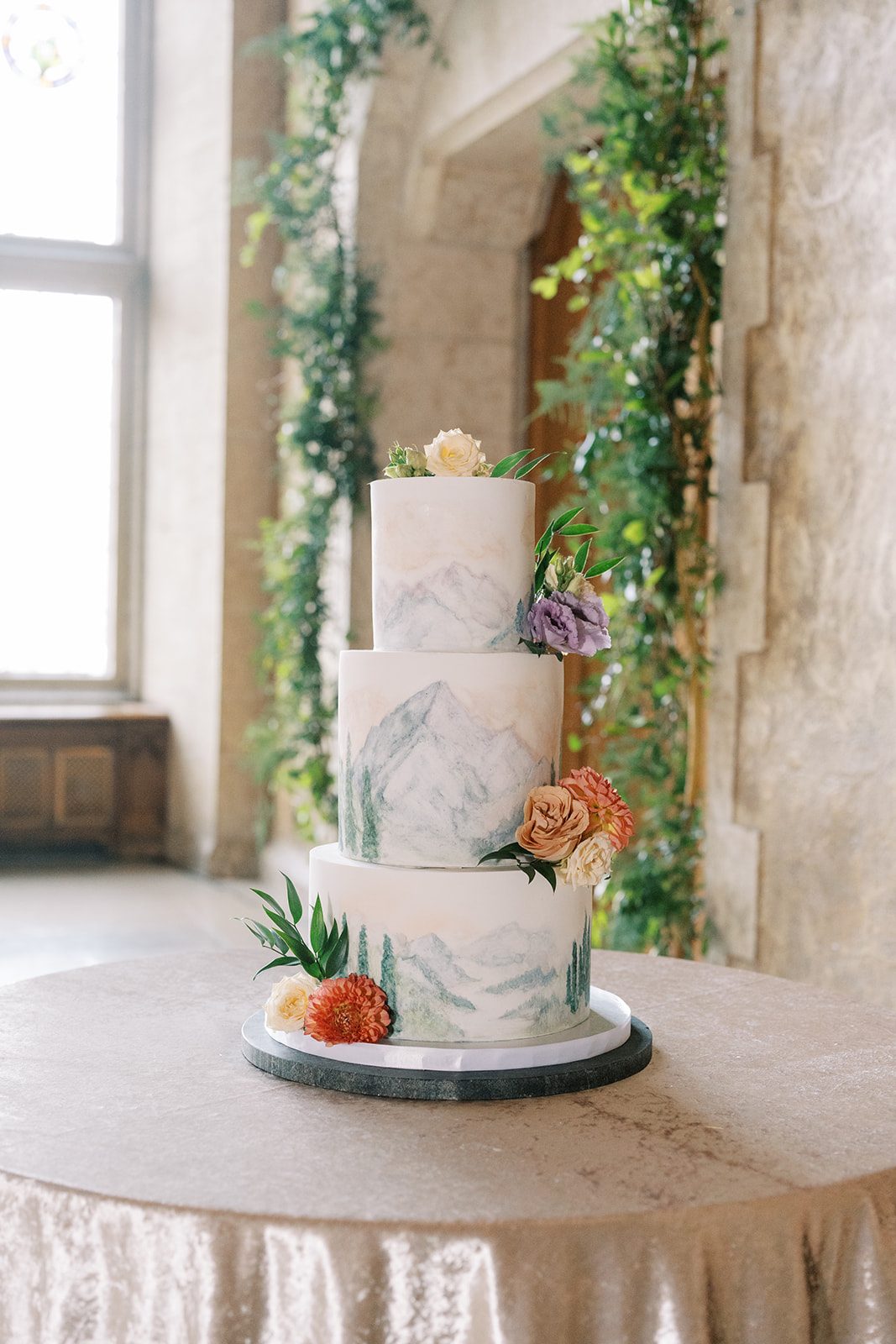 Lottie Three tier wedding cake with hand painted mountain scenery and delicate garden inspired floral accents.